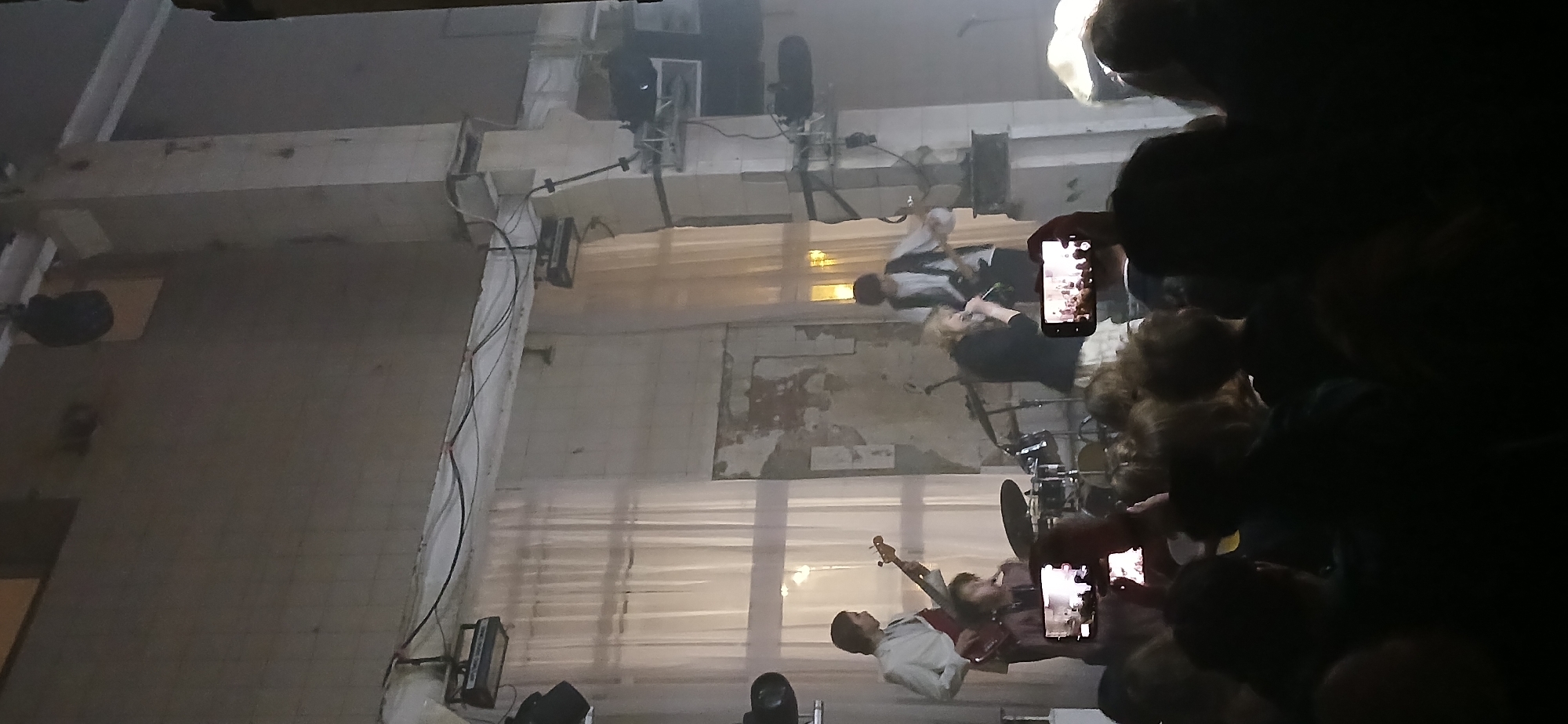 This medium shot captures a live music performance from the perspective of an audience member. The scene is an indoor concert venue with an industrial aesthetic, featuring bare concrete walls, exposed beams, and visible conduits and cables. Large, light-colored drapes or screens form a backdrop behind the performers.
On stage, at least three musicians are performing. In the center, a person with light hair, wearing a dark top, sings into a microphone. To their left, a musician in a white shirt and dark pants is visible, possibly playing a bass guitar. Another musician, dressed in a light-colored top, stands further back, and a drum set is partially visible, suggesting a drummer is present. Various stage lights and speakers are set up around the performance area, along with cabling and rigging.
In the dark foreground, the heads and shoulders of numerous audience members are silhouetted. Many attendees are holding up illuminated smartphones, actively recording the concert or taking photos of the performers. The overall lighting suggests an evening or nighttime event, with artificial stage lights illuminating the band and casting the audience in shadow. No discernible text is visible on the walls or equipment. The venue's raw, unfinished appearance suggests a repurposed industrial space, contributing to a modern or underground concert vibe.