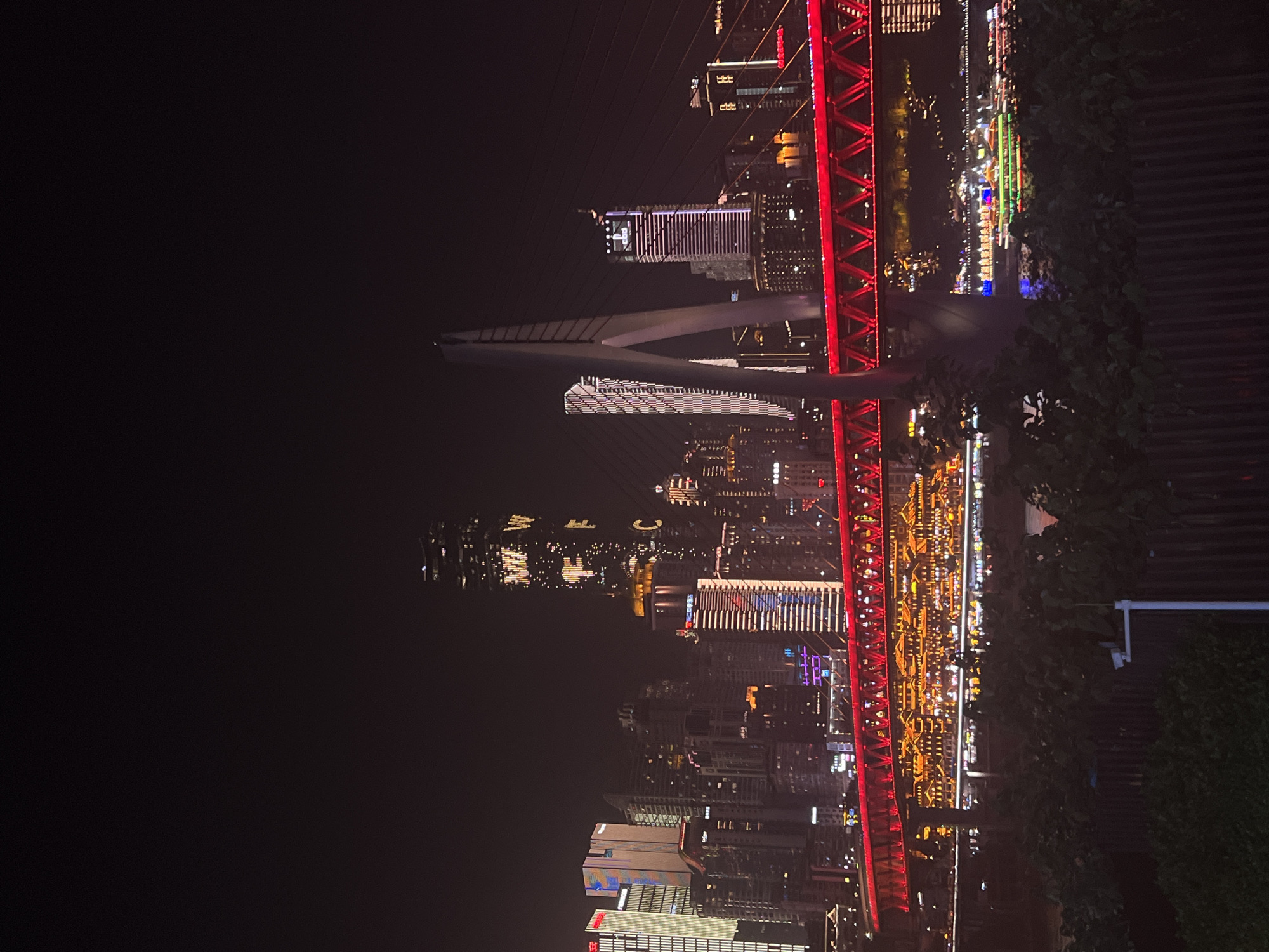The image captures a nighttime view of the cityscape of Jiangbeicheng Subdistrict, Chongqing, China. A prominent suspension bridge, illuminated with bright red lights in a zigzag pattern, spans across the river in the foreground. On the bridge, a modern architectural element, likely a support tower, is visible. Behind the bridge, the city skyline is ablaze with lights from numerous skyscrapers. One exceptionally tall building displays large illuminated letters that appear to read "WF C". Another building features a digital clock displaying "20:30". Traditional-looking buildings with golden illuminated roofs are situated along the riverfront, below the suspension bridge, reflecting in the water. Several illuminated boats are docked or moving on the river, with at least one brightly lit with green and red lights. The scene is framed by dark foliage in the very bottom of the image. The sky is dark and clear, with no visible celestial bodies. The overall impression is a vibrant, modern metropolis at night.