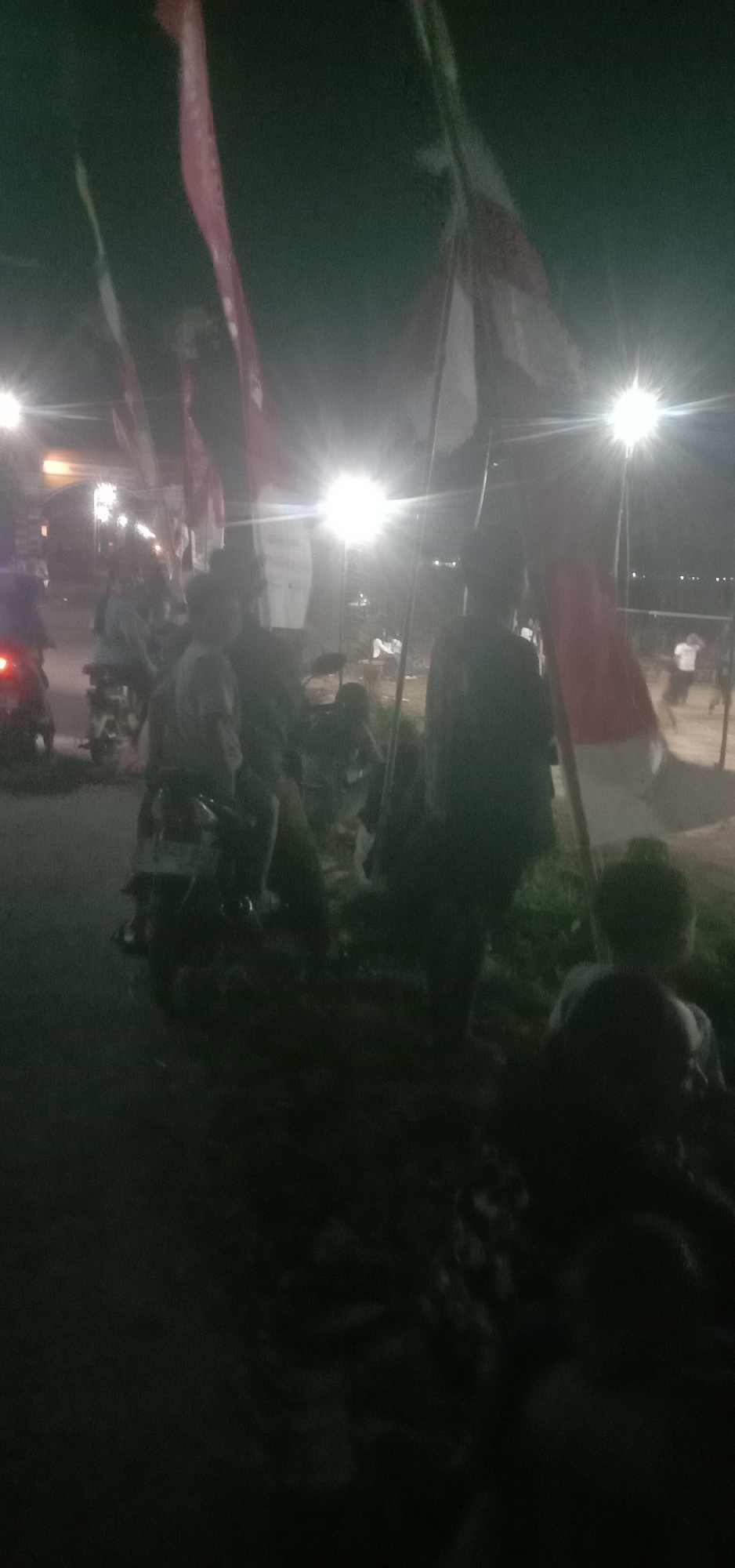 At night in Banjar Dowo, Indonesia, several individuals are gathered outdoors near a street. The scene is illuminated by artificial lights, possibly streetlights or floodlights, creating bright spots and lens flares in the image. In the foreground, a person is visible from the back, sitting or standing amongst foliage. Further back, several other people are seated or standing around motorcycles. Two motorcycles are clearly visible, one with its taillight illuminated. Multiple Indonesian flags, predominantly red and white, are displayed on poles, some appearing to flutter in the breeze. The background suggests an open area, potentially a field or dusty ground, with some structures or buildings dimly visible in the distance. The overall atmosphere is somewhat dark and blurry, indicating low light conditions and a potentially unsteady camera.