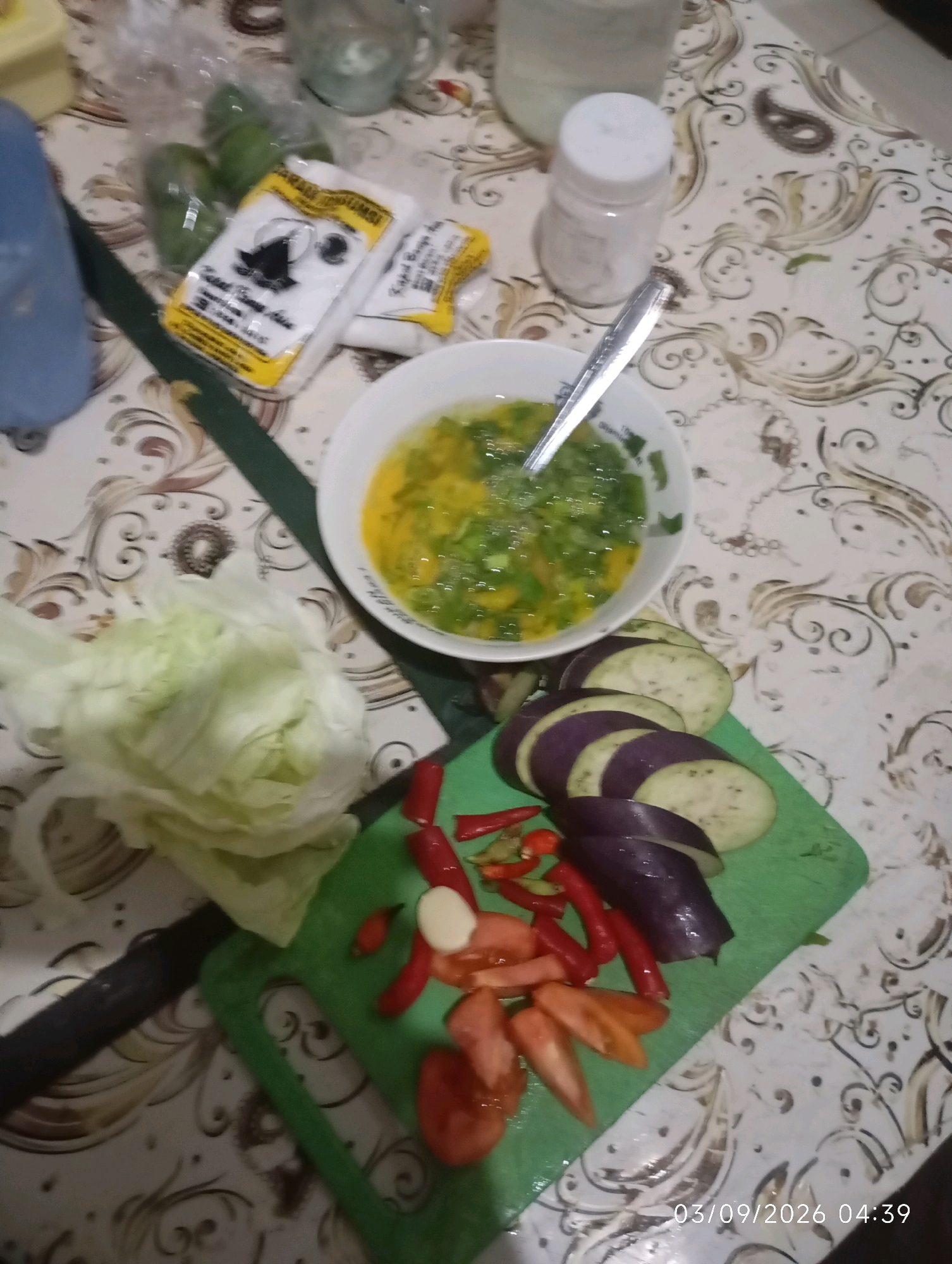 The image shows a close-up, top-down view of various ingredients laid out on a patterned tablecloth, likely in a kitchen setting in Klecorejo, Indonesia. In the center of the frame, a white bowl contains a yellow liquid with green chopped ingredients, possibly an omelet or soup base, with a spoon resting inside. To the right of the bowl, a white shaker with a silver lid is visible. In the foreground, a green cutting board displays chopped vegetables including sliced eggplant, tomatoes, red chilies, and a clove of garlic. To the left of the cutting board, a head of lettuce is partially visible. In the upper left of the image, several green fruits, possibly limes, are in a plastic bag, next to a package with yellow and white labeling. The overall scene suggests food preparation.