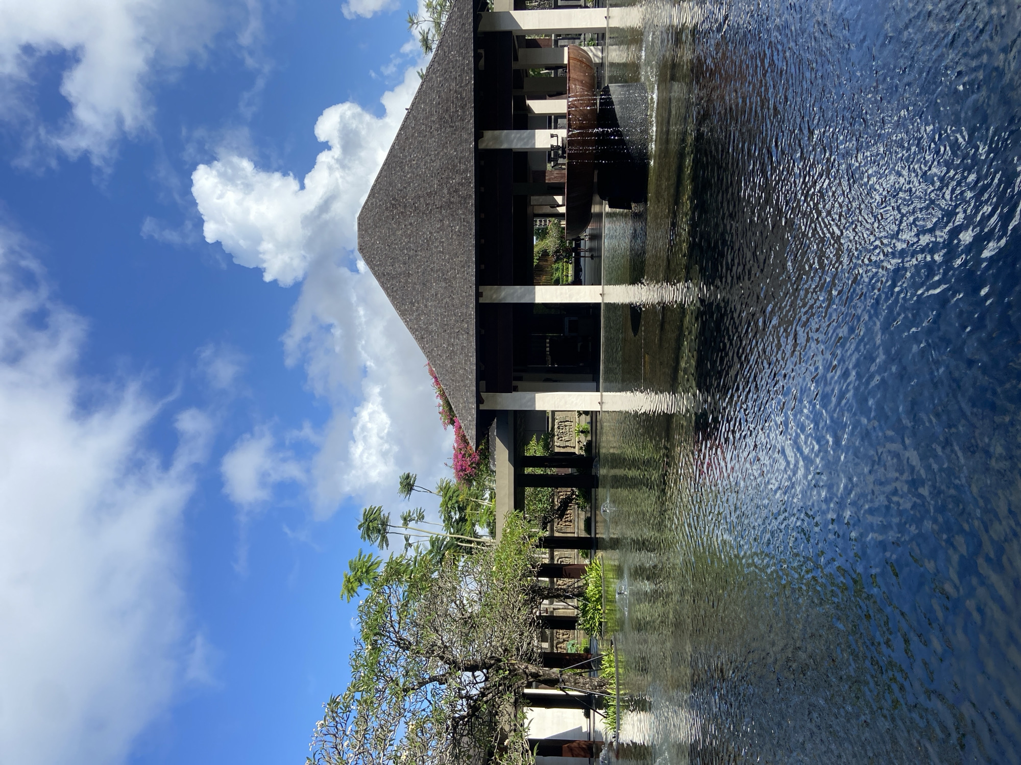 The photograph depicts a structure with a thatched roof reflected in a body of water, likely a pond or pool. The structure is supported by columns and features decorative elements. A tree with bare branches stands in the foreground, partially obscuring the view. In the background, palm trees and other greenery are visible. The sky is blue with scattered clouds. The location is inferred to be in Kuta Selatan, Indonesia.