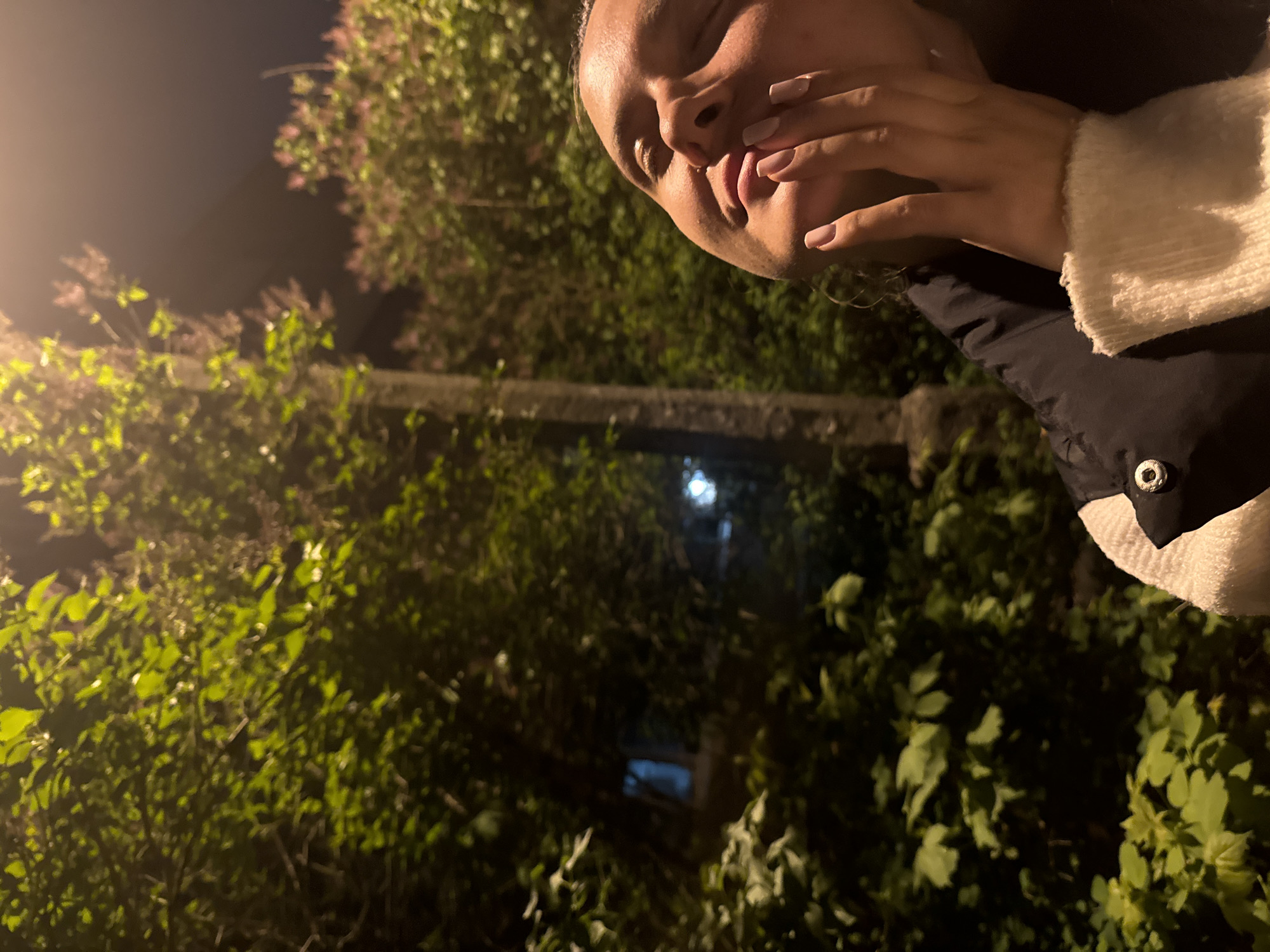 A woman is visible from the cheekbones up, turned to the right and with her mouth covered by her hand. Her complexion appears tan, and she has manicured fingernails painted in a neutral tone. She's wearing a black coat with a white sweater underneath. The background appears to be a nighttime scene with abundant vegetation, possibly a garden or park. There are green leaves and possibly some blooming purple flowers. There are other buildings visible, seemingly through a gap in the greenery. Based on the location information, the image might have been taken in a park or green area in the city of Vniissok, Russia at night.