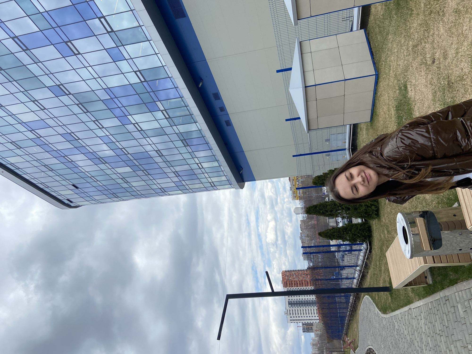 A young woman is captured in a daylight outdoor shot, smiling faintly at the viewer. She has long brown hair and is wearing a brown shiny puffer jacket.
The scene depicts an urban or suburban environment, likely a public park or walkway adjacent to modern buildings. In the foreground, a paved path with light-colored pavers curves towards the background. To the right of the path, a contemporary outdoor seating structure or bench, made of light-colored wood with metallic elements, is visible. Beyond this, a grassy area features small evergreen trees and a fence. Further back on the grass, a few small, light-colored, cube-shaped structures with blue bases are scattered.
Dominating the left side of the frame is a large, modern building with a striking facade composed of numerous blue and light blue glass panels, reflecting the sky. Above and to the right of this, another building section features a long, prominent blue horizontal stripe. The sky is overcast, filled with various shades of grey and white clouds. In the far background, several taller residential buildings and other urban structures can be seen, partially obscured by fences and trees. A black, angular street light stands out in the lower mid-ground.
The weather appears cool but bright, typical of an overcast day. No specific activities beyond the woman posing for the photograph are evident. No visible text is present in the image. The setting suggests a modern cityscape in Izhevsk, Russia.
