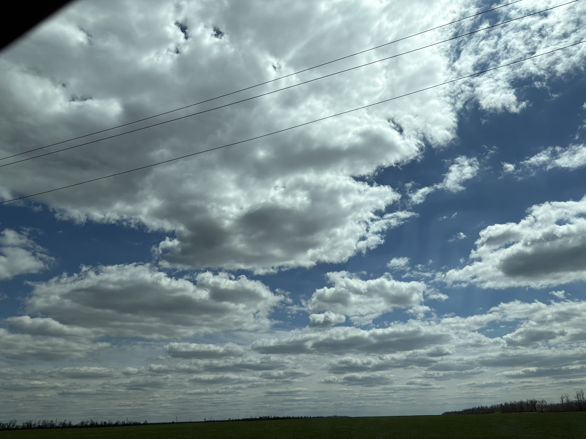 The image shows a view of the sky with numerous clouds, interspersed with blue patches. There are utility lines visible in the upper part of the frame. The horizon consists of a green field, with a few trees visible in the distance on the right side of the frame. The location is consistent with an open area in or near Margaritovo, Russia. No people are visible.