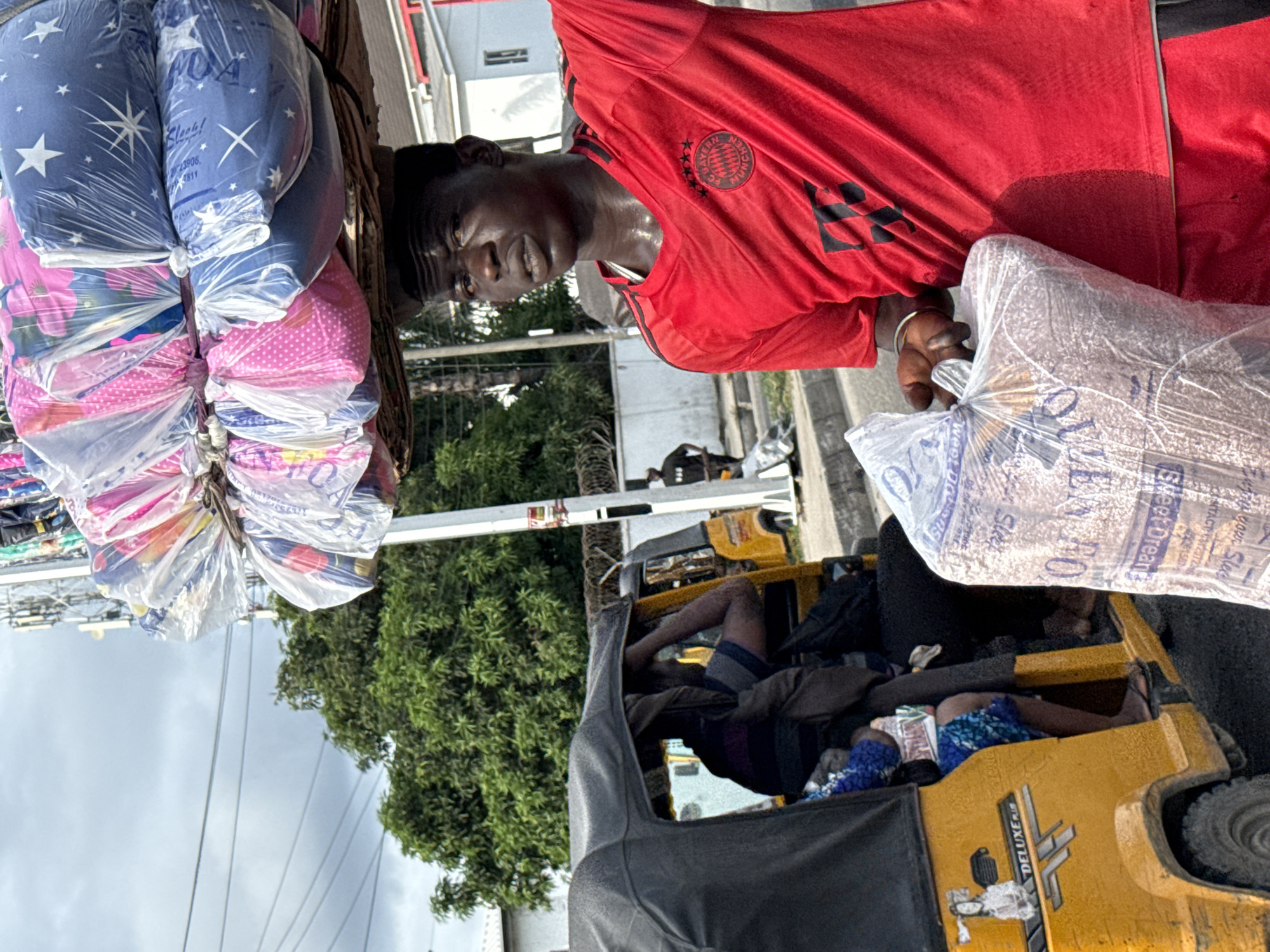 This image captures a vibrant street scene, likely in Ado, Nigeria, centered around a mobile vendor. The main subject is a dark-skinned man, seen from the chest up, wearing a red short-sleeved jersey with the "FC BAYERN MÜNCHEN" logo visible on his left chest. His face shows signs of perspiration, suggesting warm weather or exertion, and he has a neutral to slightly strained expression, looking towards the viewer.
He is burdened with merchandise: a tall stack of bundled textiles or bedding, wrapped in clear plastic, is carried on his head. These bundles display various patterns, including blue with white stars, pink with white polka dots, and floral designs. Some visible text on these bundles includes "Klook!", "223906", and "4811". In his left hand, he carries a large, clear plastic bag filled with what appears to be light-colored grains or small particles, possibly dry food items. Text on this bag includes "your Sleep", "Sweet Dream", and "CONTACT 2372".
In the background and to his right, a yellow three-wheeled auto-rickshaw, branded "DELUXE", is visible. Inside the vehicle, several individuals are present, some appearing to be resting. The setting includes green foliage from trees, a partly cloudy sky, and hints of distant buildings, typical of an urban or semi-urban environment. The man's activity suggests he is a street hawker, either walking or briefly paused, selling his goods in what appears to be a busy area.