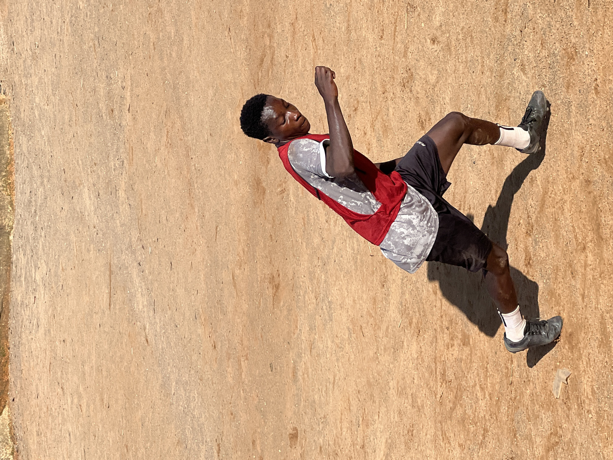 A young Black male wearing a red training bib over a grey and white patterned t-shirt and black shorts is captured in mid-stride on a dusty, orange-brown dirt field. He is wearing white socks and black cleats. His facial expression is focused as he extends his right arm forward, suggesting he is in motion, possibly playing or practicing a sport. A long shadow stretches behind him to his left, indicating the sun is positioned to his right. The environment appears to be an outdoor, likely urban, setting in Jos, Nigeria, with the ground showing signs of wear and sparse debris.