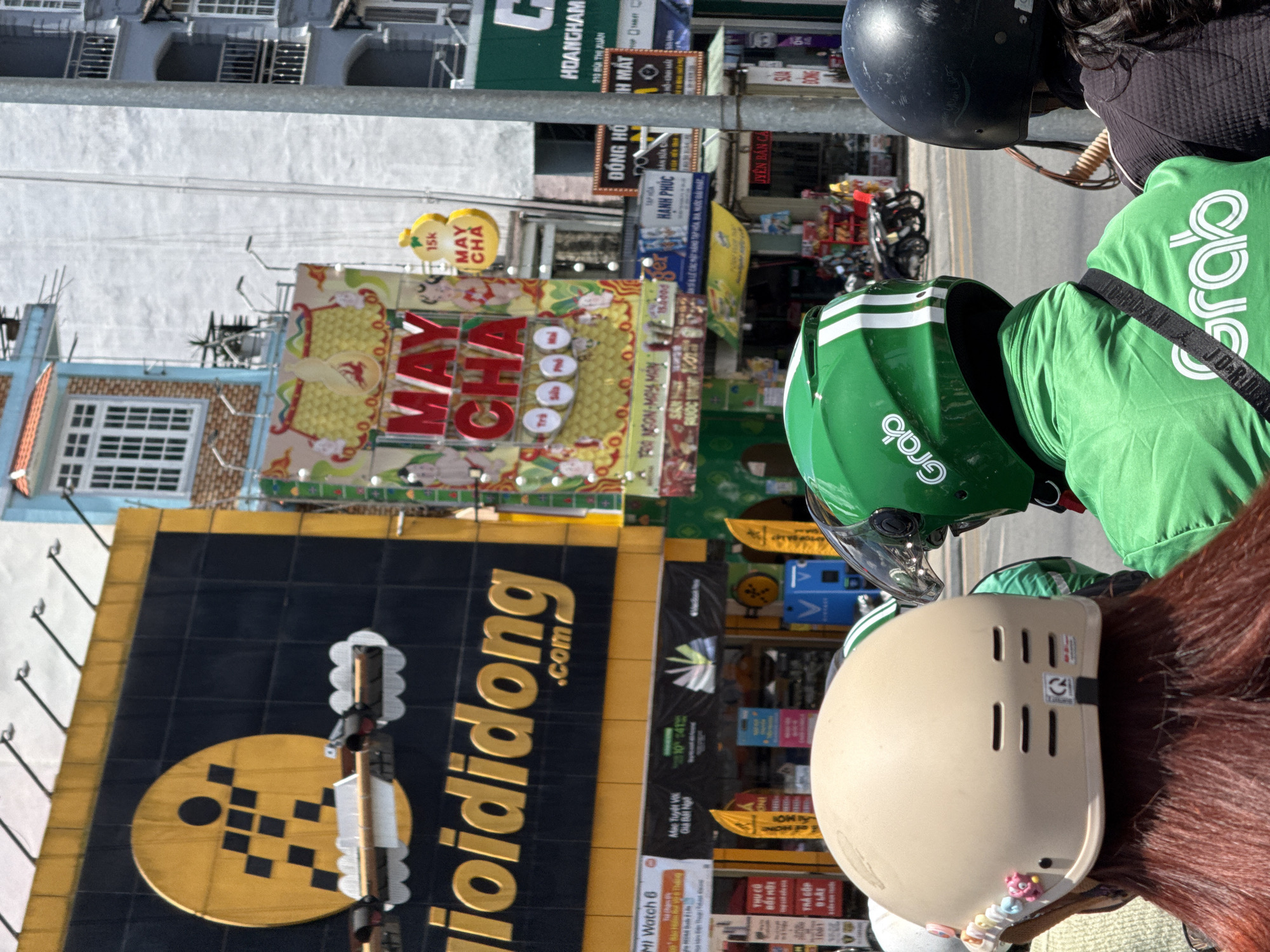 The image depicts a street scene in Da Lat, Vietnam. A large black and gold sign for 'ioididong.com' is prominent on the left, advertising what seems to be a tech store. Nearby, there is a smaller sign for 'MI Watch 6.' In the background, there are various advertisements for goods and food, including a bright sign for 'MAY CHA' with a price of 15k (presumably Vietnamese Dong). On the right side of the frame, the back of a person wearing a green Grab delivery jacket and helmet is visible. Another person wearing a tan helmet with decorations is sitting behind the Grab driver. There are also other people visible with black helmets. Some traffic can be seen in the background.
