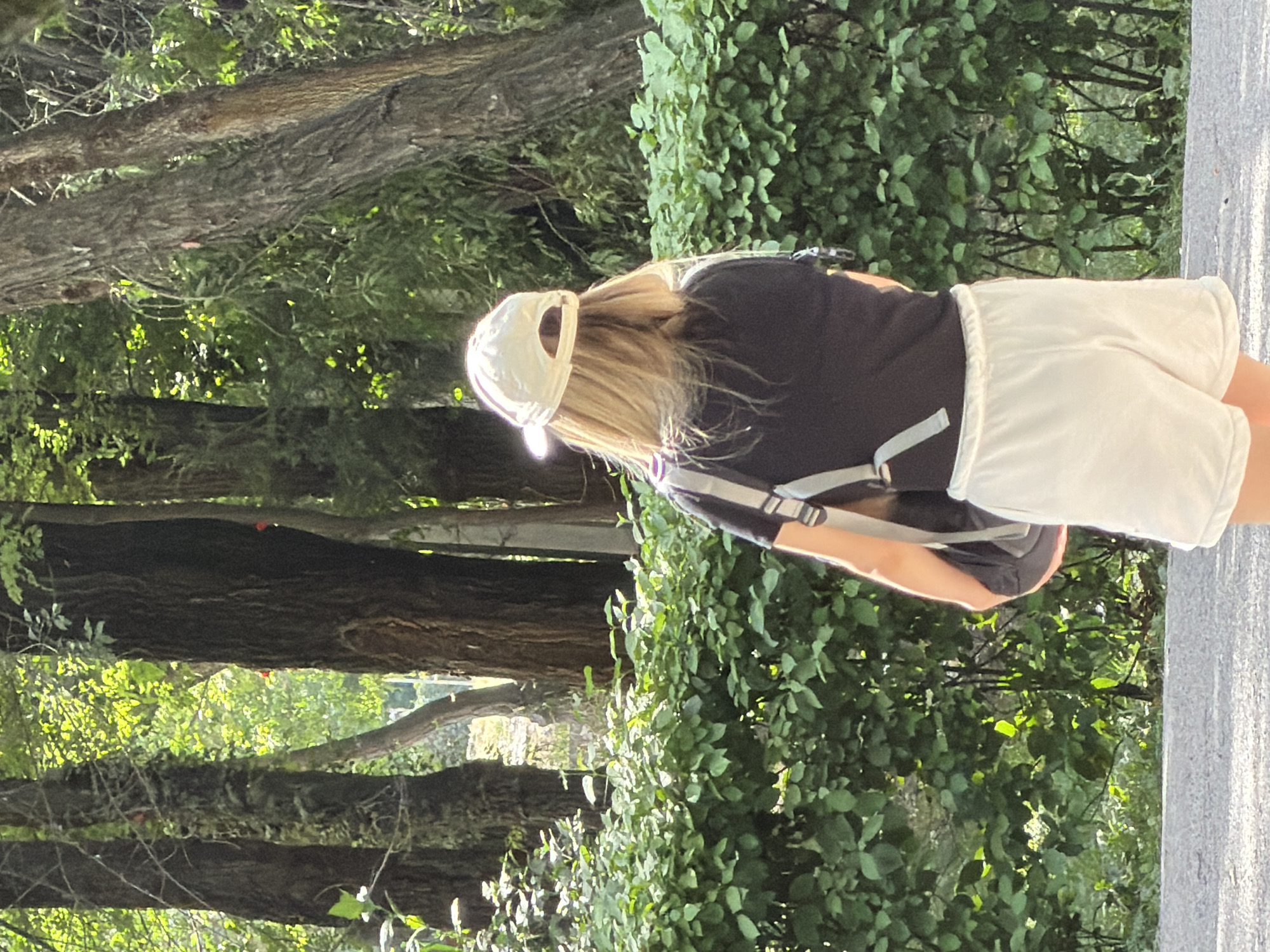 A person with blonde hair, wearing a black t-shirt, white shorts, and a white baseball cap, stands facing away from the camera, looking towards a dense green hedge. They are wearing a backpack with grey straps. The background consists of tall trees with dappled sunlight filtering through their leaves. The ground in front of the hedge is paved.
