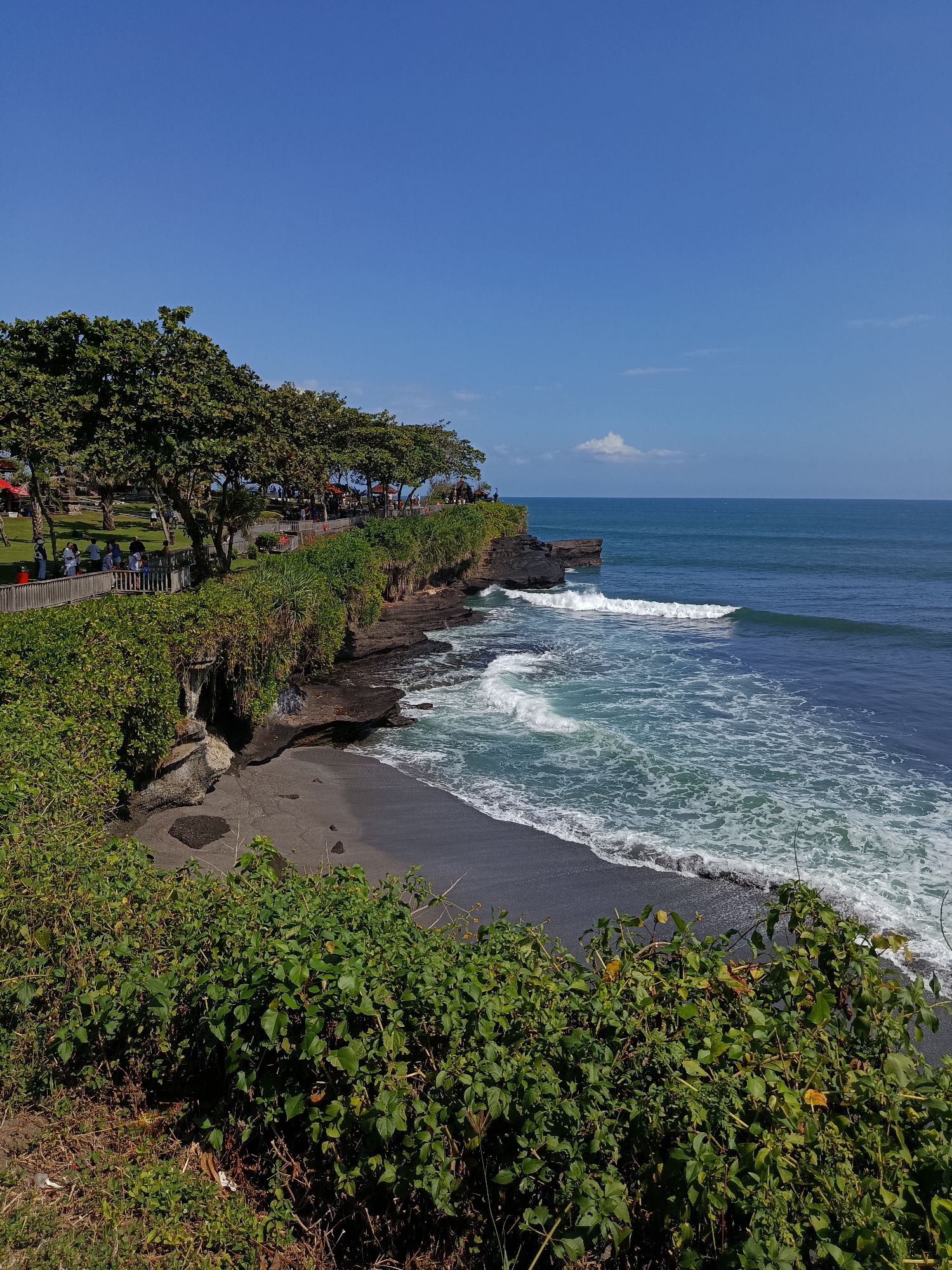 This image captures a scenic coastal view with lush green foliage and a dark sandy beach meeting the ocean. The scene depicts a cliffside area adorned with dense vegetation, trees, and a wooden walkway. Several individuals are visible walking along the pathway, observing the seascape. The ocean displays moderate waves breaking on the shore, with clear blue skies above. The location is identified as Tanah Lot Temple, which is situated in the village of Beraban, Tabanan Regency, Bali, Indonesia, not in Kediri.