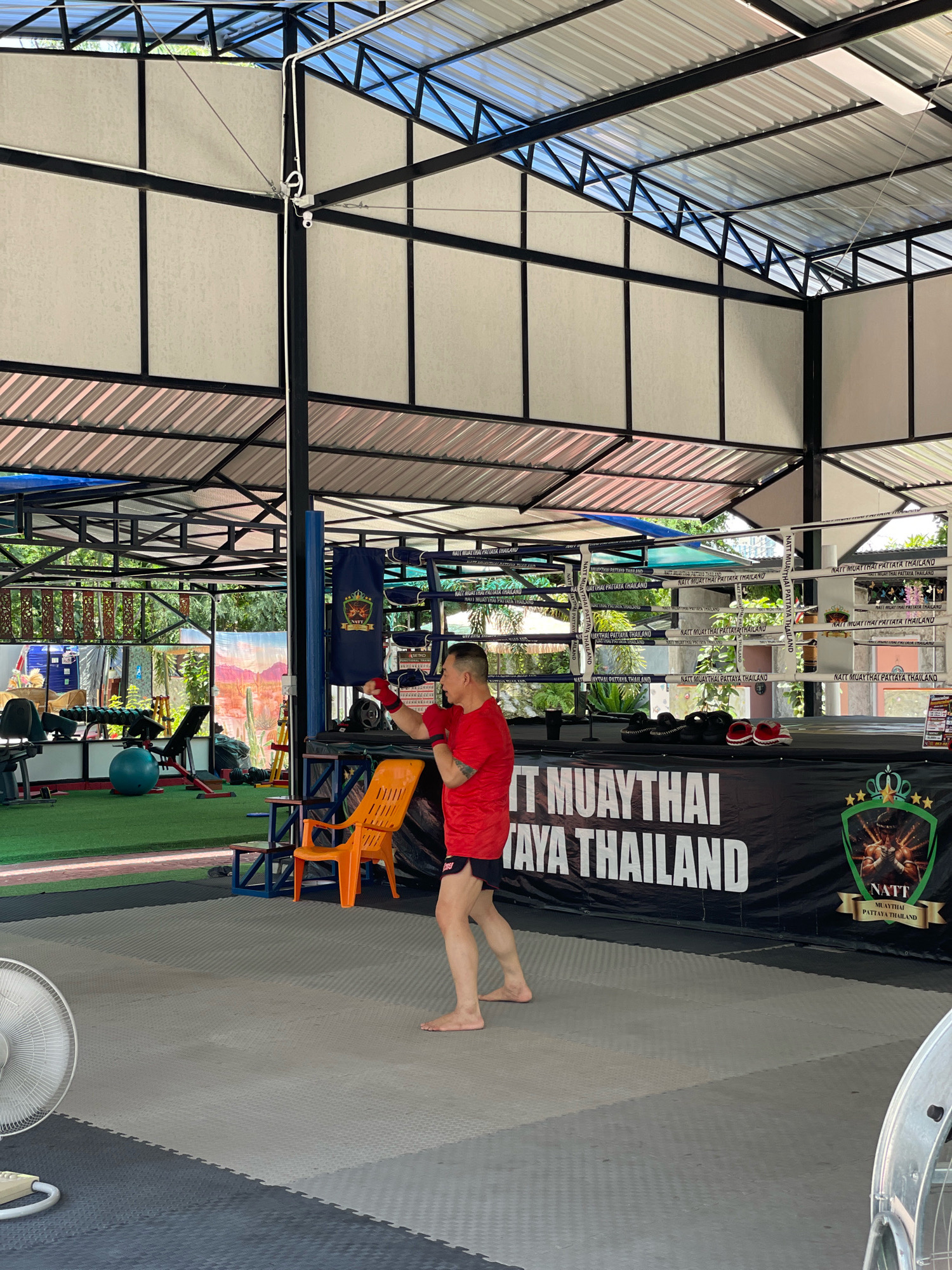 A man in a red shirt and black shorts is practicing Muay Thai in an open-air gym in Pattaya City, Thailand. The gym has a metal roof supported by a black metal frame, with corrugated metal on the outer edges and translucent panels in the upper sections. A boxing ring is set up in the background, with a black banner behind it that reads "NATT MUAYTHAI PATTAYA THAILAND" and displays the gym's logo. To the left of the ring, there are various exercise machines, a bright blue exercise ball, and an orange chair. The man is barefoot and wearing red hand wraps. He is in a defensive stance, with his left hand extended and his right fist held near his face. A large white fan is visible in the foreground to the left. The ground is covered with grey and black puzzle mats. The environment appears to be outdoors, with greenery visible through the open sides of the gym.