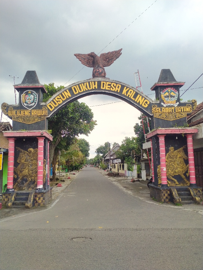 This image presents a welcoming archway marking the entrance to a village or hamlet in what appears to be a rural or semi-urban setting in Kecamatan Kebakkramat, Indonesia. The archway, predominantly black and gold, spans a paved road that extends straight into the distance, flanked by residential houses and trees under an overcast sky.
A large, brown eagle sculpture with outstretched wings is prominently perched atop the center of the arch. Bold gold letters on the arch clearly state "DUSUN DUKUH DESA KALING." The arch is supported by two decorative pillars, also in black and gold with pinkish-red accents. The left pillar features an emblem and the Javanese welcome phrase "WILUJENG RAWUH!", while the right pillar displays a similar emblem and the Indonesian welcome "SELAMAT DATANG." Both pillars are adorned with golden bas-reliefs depicting dynamic figures, seemingly warriors, on horseback.
Beyond the arch, the asphalt road is visible, with modest, single-story homes lining both sides. The houses exhibit typical Indonesian architectural styles, with tiled roofs and some greenery. Numerous trees provide shade along the street. In the far distance, a few small figures can be discerned, suggesting people going about their daily routines. The sky is uniformly overcast, indicating either a cloudy day, early morning, or late afternoon, with diffused lighting across the scene. Power lines are visible stretching across the top of the frame.