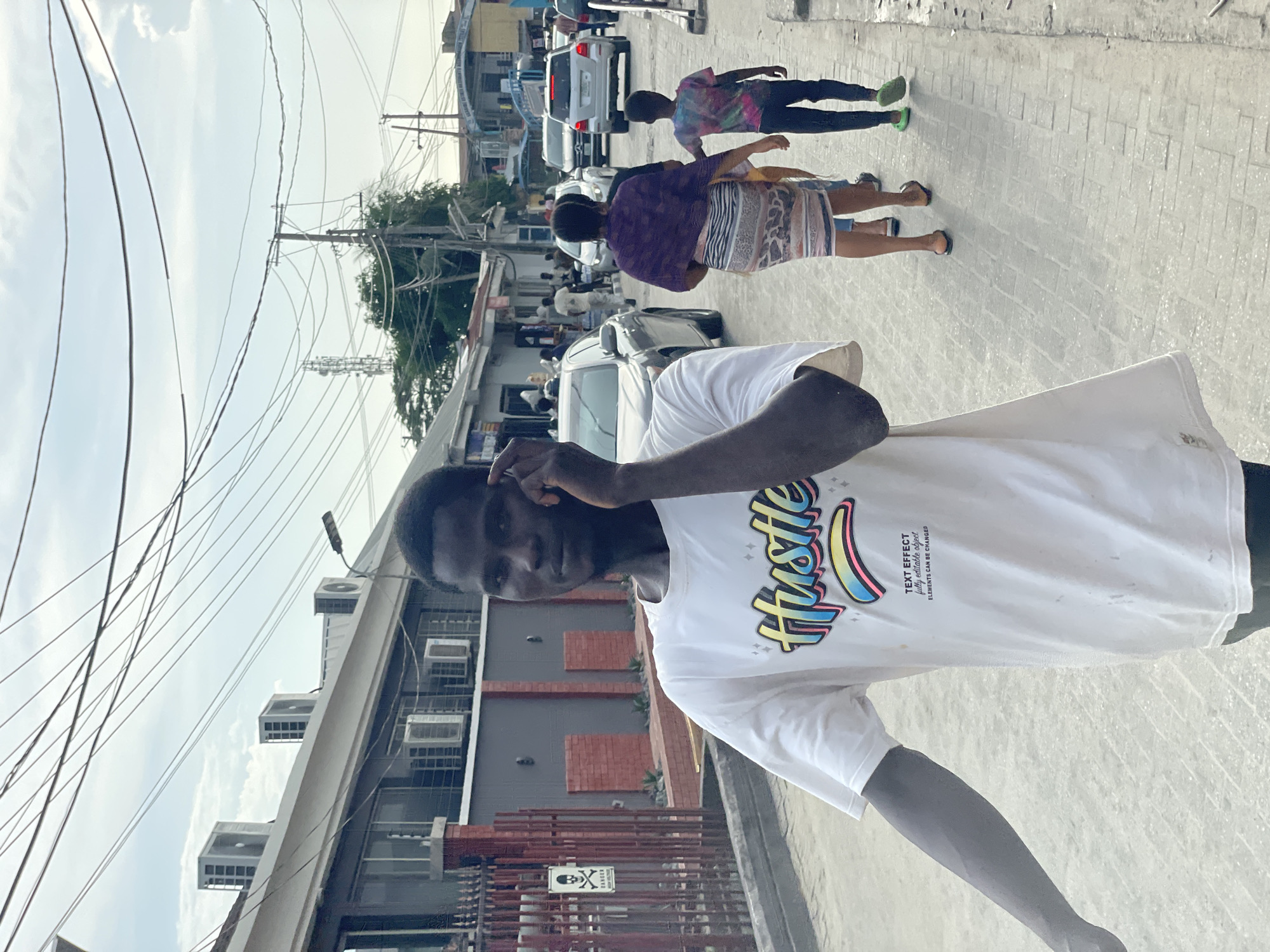 The image captures a vibrant street scene, appearing rotated 90 degrees counter-clockwise from its natural orientation. In the immediate foreground, a young man with dark skin looks directly at the camera, his right hand near his head and his left arm extended downwards. He wears a white t-shirt featuring the word "Hustle" in colorful, stylized lettering. Below "Hustle," smaller text reads: "TEXT EFFECT fully editable object ELEMENTS CAN BE CHANGED."
Behind the main subject, a paved street extends into the distance, bustling with activity. Several people are visible walking; one person wears a purple top and a patterned skirt or shorts, while another is in a tie-dye shirt and dark trousers with distinctive bright green shoes. Various vehicles, including cars and SUVs, are present on the street, some parked and others seemingly in motion.
Buildings line both sides of the street. On the left, a multi-story building features several external air conditioning units. Utility poles and a dense network of overhead power lines crisscross against a bright, cloudy sky, suggesting a clear day. Near a fence or gate of one building, a sign with a skull and crossbones symbol, commonly denoting "DANGER," is visible. The overall setting appears to be an urban or semi-urban area in Iranla, Nigeria, characterized by local architecture and street life under daylight conditions.
