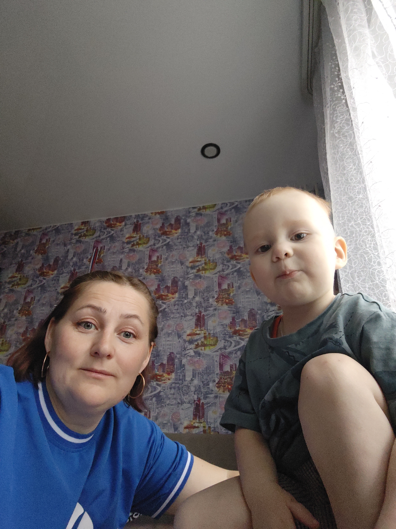 The image depicts a woman and a young child. The woman, fair-skinned with light brown hair pulled back, wears a blue t-shirt with white stripes. She is wearing earrings, and her expression is neutral. The child, also fair-skinned with reddish-blonde hair, wears a grey-green t-shirt. The child has a slightly puckered expression. They appear to be indoors, with wallpaper visible in the background featuring abstract patterns and cityscape elements. A white curtain is visible near a window in the upper right corner of the image. The location is Markovskii, Russia, evidenced by the cityscape wallpaper. The camera angle is from above, looking down at the subjects.