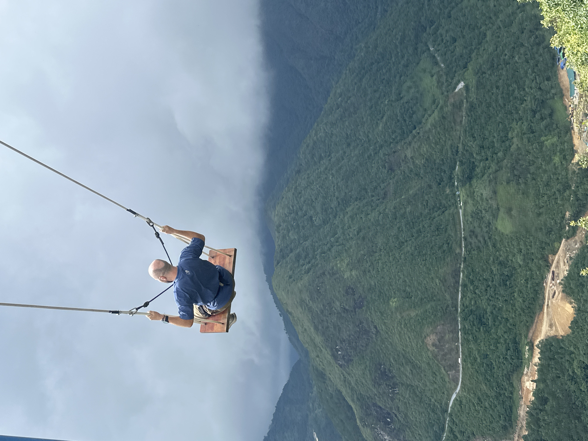 A man sits on a wooden swing suspended over a steep, mountainous landscape in Binh Lu, Vietnam. The man is bald, wearing a blue short-sleeved shirt, beige shorts, and shoes. He grips the swing's ropes with both hands. Below him, a deep valley is filled with lush green trees and obscured by clouds. A road winds through the landscape. The sky above is overcast.