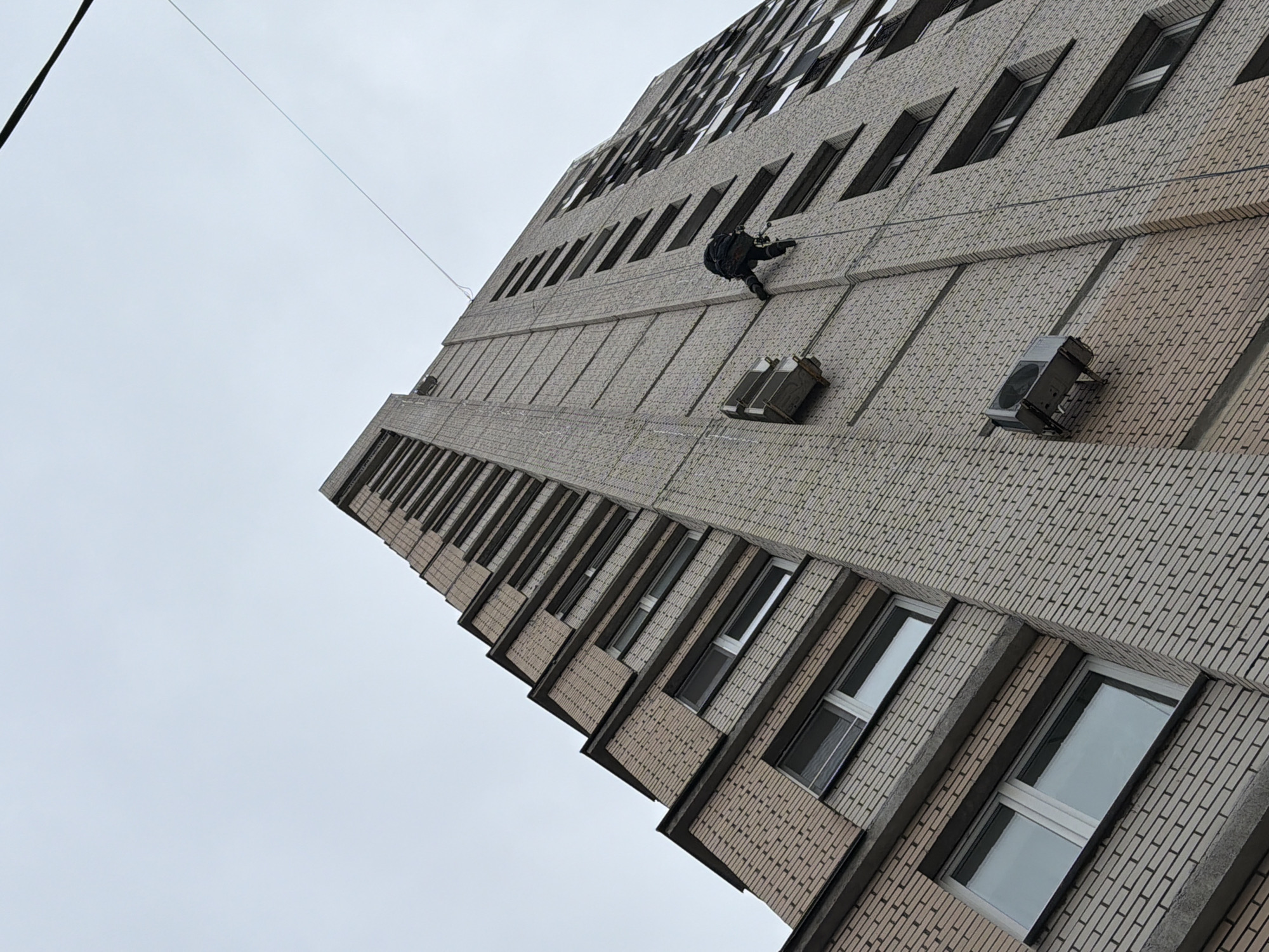 A low-angle shot captures a tall building in Saint Petersburg, Russia, with a worker rappelling down the exterior. The building has a light-brick facade with repetitive window patterns and jutting, horizontal ledges. The worker is equipped with rappelling gear, including ropes and a harness. Mounted air conditioning units are spaced along the building's facade. An electric power cable extends across the top-left corner of the frame against a predominantly overcast, pale sky.