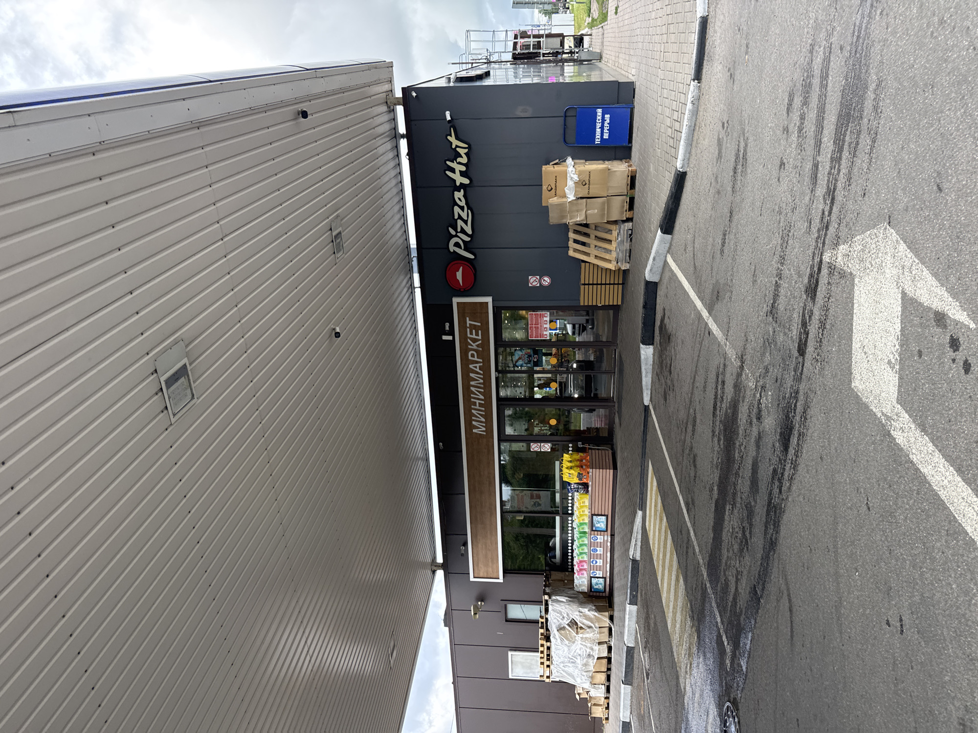 The image shows the exterior of a combined mini-market and Pizza Hut restaurant, likely located at a gas station in Saint Petersburg, Russia. The building has a dark gray facade with a prominent Pizza Hut logo and a sign that reads "МИНИМАРКЕТ" (Minimarket) in Russian. To the left of the entrance, there are stacks of goods, including pallets wrapped in plastic and cardboard boxes, suggesting deliveries or stock. In front of the minimarket, there is a display of various bottled beverages. A pedestrian crossing with yellow and white stripes is visible in the foreground, leading to the building. The area appears to be a paved lot, possibly a parking area, with tire marks on the asphalt. A blue trash bin with Russian text, "ТЕХНИЧЕСКИЙ ПЕРЕРЫВ" (Technical Break), is situated to the right of the entrance. The architecture of the building, including the overhanging canopy, is characteristic of a roadside service area. The sky is overcast with clouds, indicating a daytime scene under diffused light.