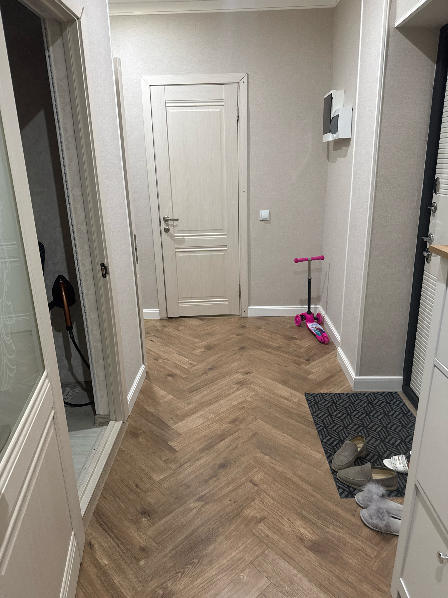 The image shows a hallway in a residential apartment in Grozny, Russia. The hallway has light-colored walls and a herringbone pattern wood floor. On the left, a partially opened door reveals a closet with an iron inside. In the center of the hallway, a closed light-colored door is visible. Further down, a pink children's scooter with characters from Frozen is parked. To the right, a door with black and white paneling is partially visible, and next to it, a white cabinet. On the floor near the cabinet, a doormat with a geometric pattern is placed, with several pairs of shoes on and around it, including gray loafers and fluffy slippers. A wall-mounted unit, possibly an intercom or a small shelf, is located on the right wall.