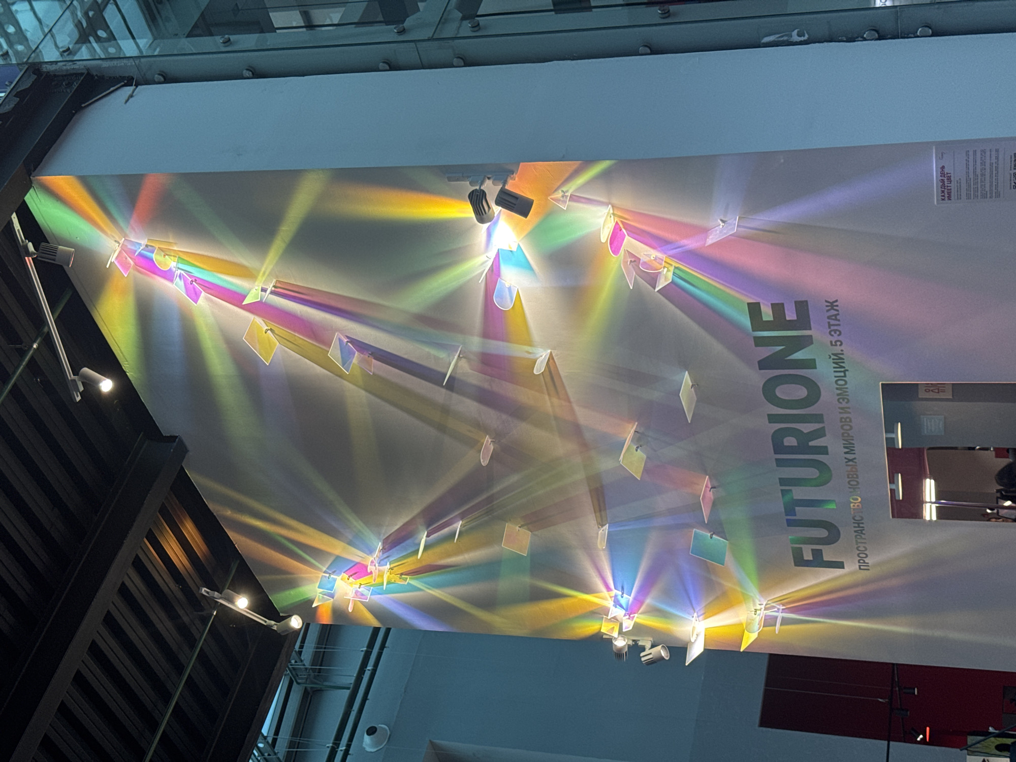 The image depicts an art installation in Saint Petersburg, Russia, likely at the "Futurio ne" exhibition as indicated by the text on the wall. The installation features numerous small, flat, square and rectangular objects mounted on a large, light-colored wall. These objects are illuminated by multiple spotlights positioned both above and to the side of the installation. The light passing through or reflecting off these objects creates a vibrant display of rainbow-colored beams that fan out across the wall. The overall effect is a dynamic and colorful light show. Below the installation, the word "FUTURIONE" is prominently displayed in large, dark green letters, followed by smaller Russian text that translates to "space of new worlds and emotions. 5th floor." The environment appears to be an indoor exhibition space, with a dark ceiling with exposed beams and track lighting visible at the top. To the right of the installation, a glass facade with metal framing suggests a modern architectural setting. No individuals are clearly identifiable in the image.