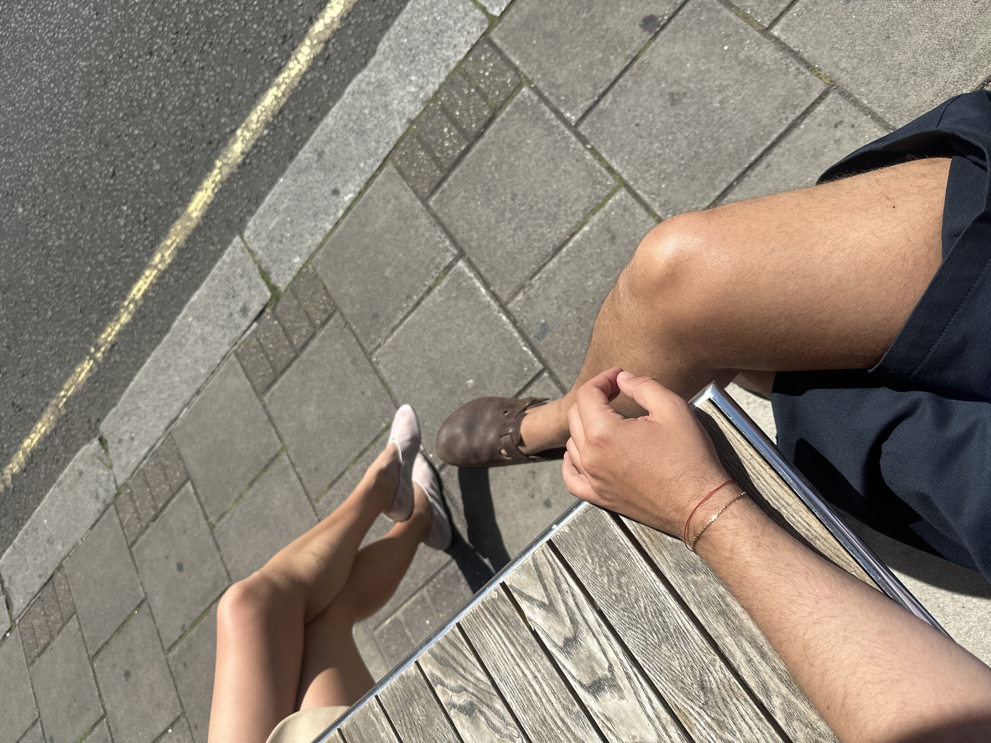 The image captures an outdoor scene on a paved area, likely a sidewalk, alongside a road marked with a yellow line. Two individuals are visible: a person with light-toned legs wearing light-colored shoes and a person with tan skin wearing brown shoes and navy blue shorts. Both appear to be seated at a wooden table with metal trim, positioned at an angle to the street. The person in shorts has a reddish bracelet on their left wrist. The location is in the city of Unknown, Unknown.