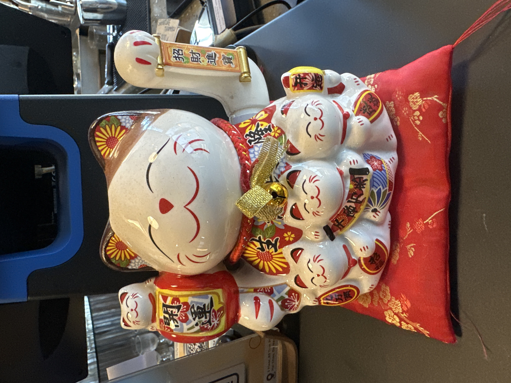 A Maneki-neko, or "lucky cat" figurine, sits on a red, gold-embroidered cushion on a dark countertop. The figurine is white with brown and yellow floral patterns on its ears. It has a red mouth and red accents on its eyes. A red collar with a gold bell is around its neck. The large cat has several smaller cats on its front. The large cat's left paw is raised and holding a scroll with Chinese characters. Its right paw is also raised, but not clearly visible. The smaller cats also have their paws raised, some holding miniature scrolls or koban coins. In the background, a black and blue piece of machinery, possibly a coffee machine, is visible, along with some stainless steel objects and glass cups. The scene appears to be indoors, possibly in a cafe or shop, given the background elements. There are no people visible in the image. The lighting suggests an indoor setting. The image quality is clear, allowing for detailed observation of the figurine and its surroundings. The overall impression is one of an ornate and decorative item displayed in a commercial or domestic setting in Đà Nẵng, Vietnam.