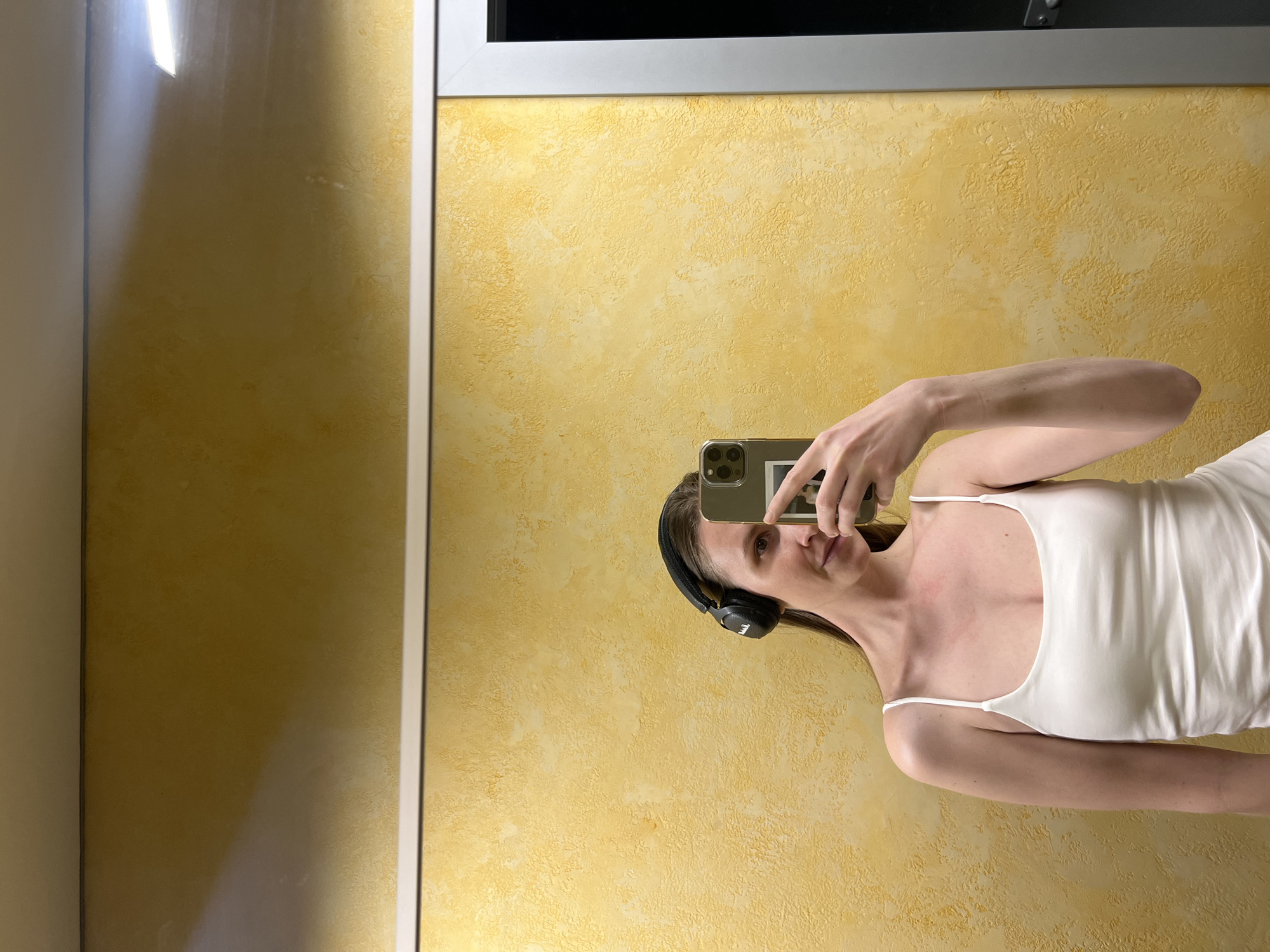 A person stands facing a mirror while taking a photo with their phone. They are wearing a white tank top and black headphones. The background is a yellow wall with a textured surface. The location is not discernible.