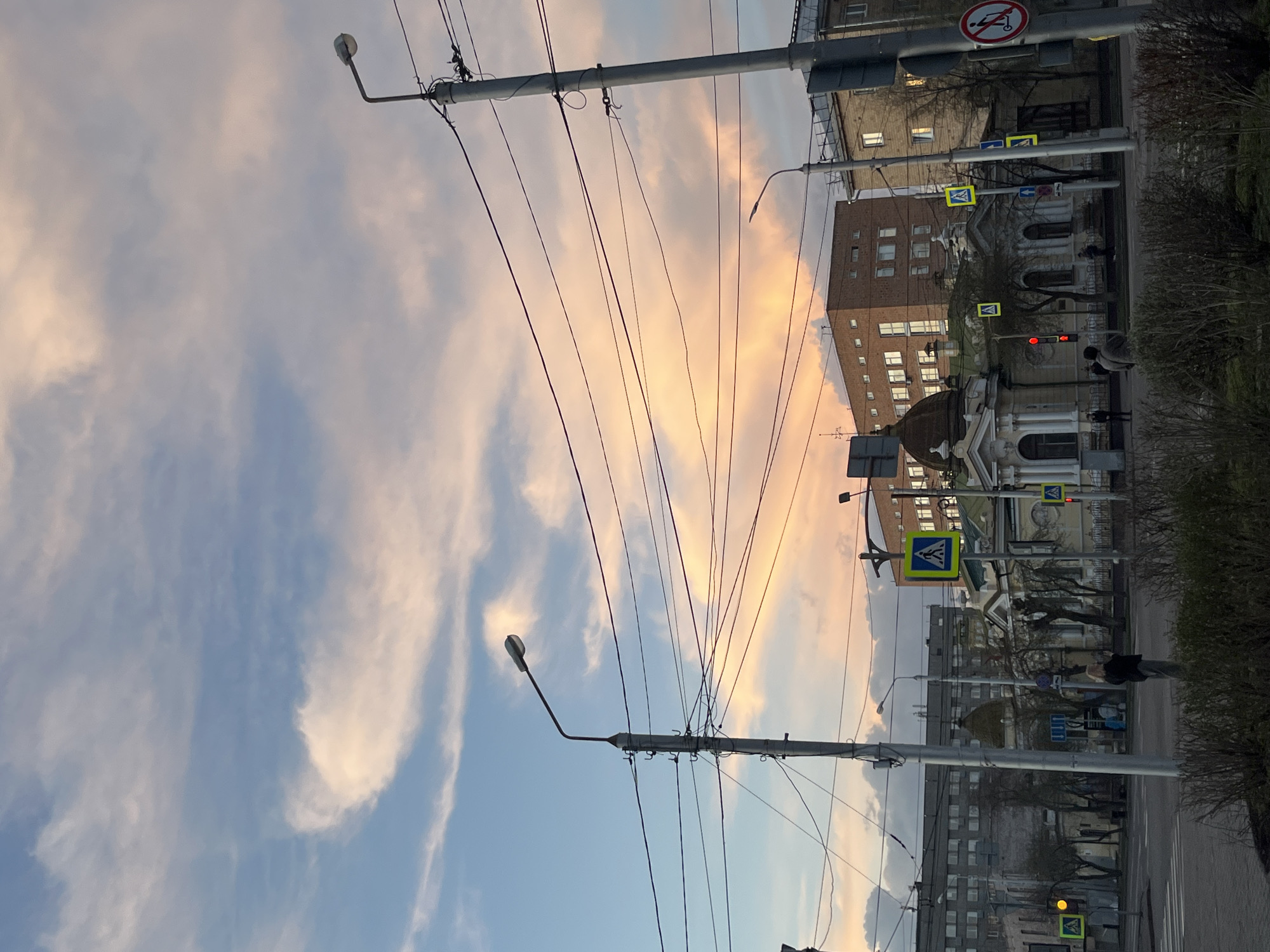 A low-angle perspective of a street scene in Krasnoyarsk, Russia, captures the intersection of urban infrastructure and the natural sky. Power lines converge from two weathered, gray utility poles, creating a dense web against the backdrop of a sky streaked with pastel hues of blue, peach, and white clouds, suggesting either sunrise or sunset. Buildings line the right side of the frame, their architectural styles varying and hinting at different periods of construction. Several traffic lights are visible, with at least one displaying a red signal. Pedestrian crossing signs are mounted on posts, indicating designated areas for pedestrians. A person in dark clothing is present on the sidewalk on the right side of the frame. Hedges or shrubs line the street, adding a touch of greenery to the urban environment. The pavement is visible as a clear surface for movement.