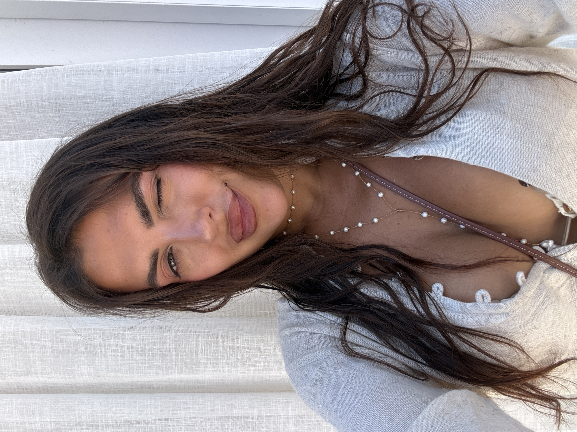 A close-up, horizontally oriented photo shows a young woman with long, dark brown hair. She is wearing a light-colored, loose-fitting top with a V-neckline and pearl-like buttons. A delicate pearl necklace is visible around her neck. Her hair is spread across the frame, framing her face and body.
The woman is looking directly at the camera with one eye open and the other winking. Her tongue is sticking out slightly, and her lips are parted. Her expression appears playful and flirtatious. She is positioned against a backdrop of white, textured curtains or blinds, which are evenly spaced and create a striped pattern. The lighting suggests it is daytime, and the overall mood is casual and relaxed. The location is unknown but could be a home, hotel room, or outdoor covered area with such window coverings. There is no visible text in the image.