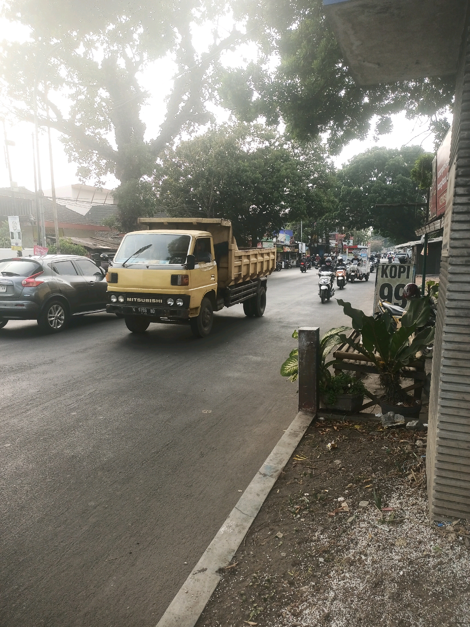 A yellow Mitsubishi dump truck is parked on the left side of a street in Malang, Indonesia. The truck is facing forward and appears to be empty. A dark gray SUV is parked behind the truck. The street is asphalt and appears to be wet, possibly from rain. There are trees and buildings on both sides of the street. The sky is overcast and hazy. In the background, there are several motorcycles and other vehicles on the road, and people can be seen riding them. A sign that says "KOPI 99" is visible on the right side of the street, near some potted plants.