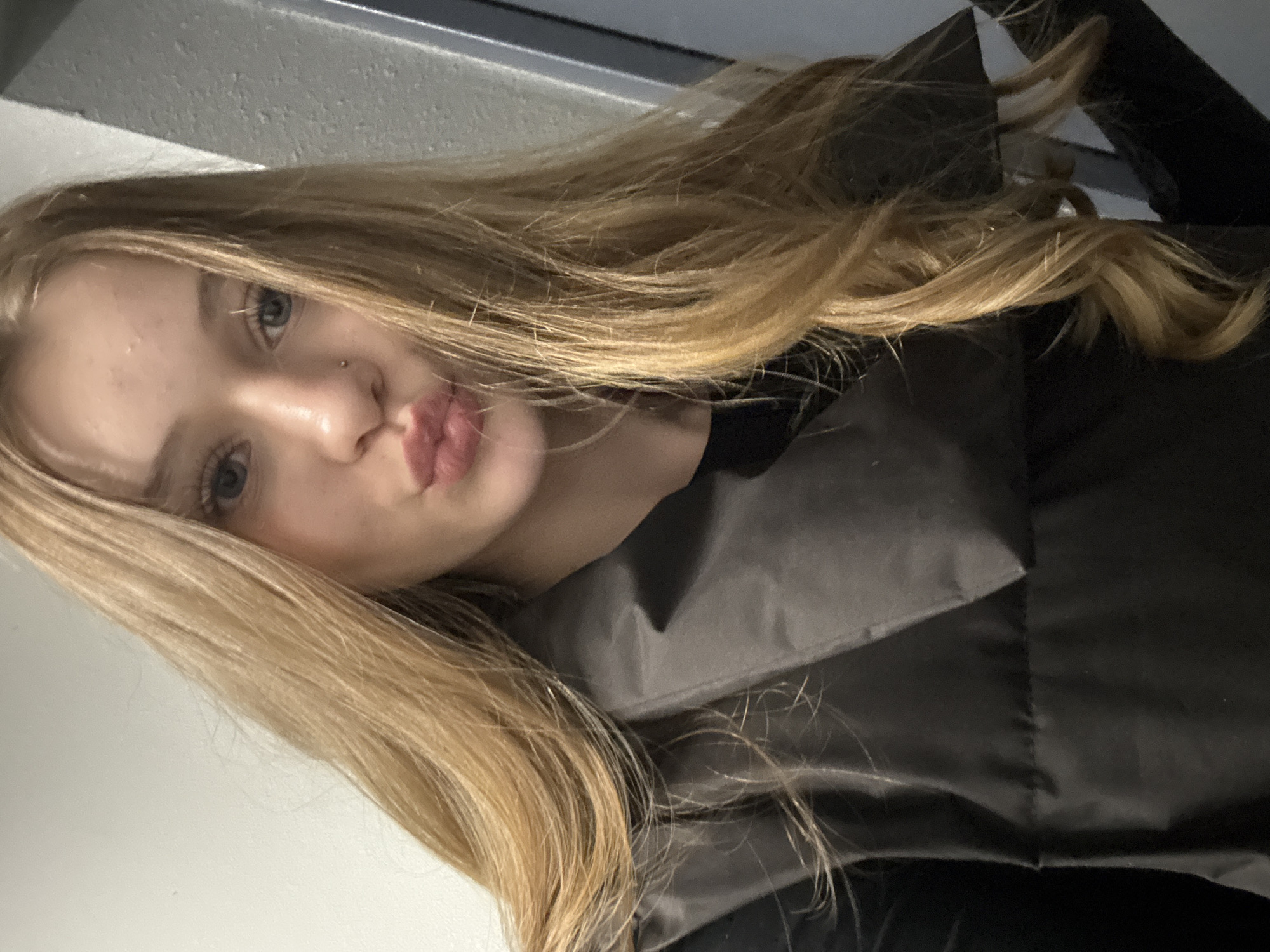 A young, fair-skinned woman with long, blonde hair is facing the camera with slightly pursed lips. She has light eyes and a stud piercing in her nose. She is wearing a dark-colored vest or jacket. The background consists of a white wall and a gray metallic or concrete structure. There is no indication that this photo was taken in Yekaterinburg, Russia.