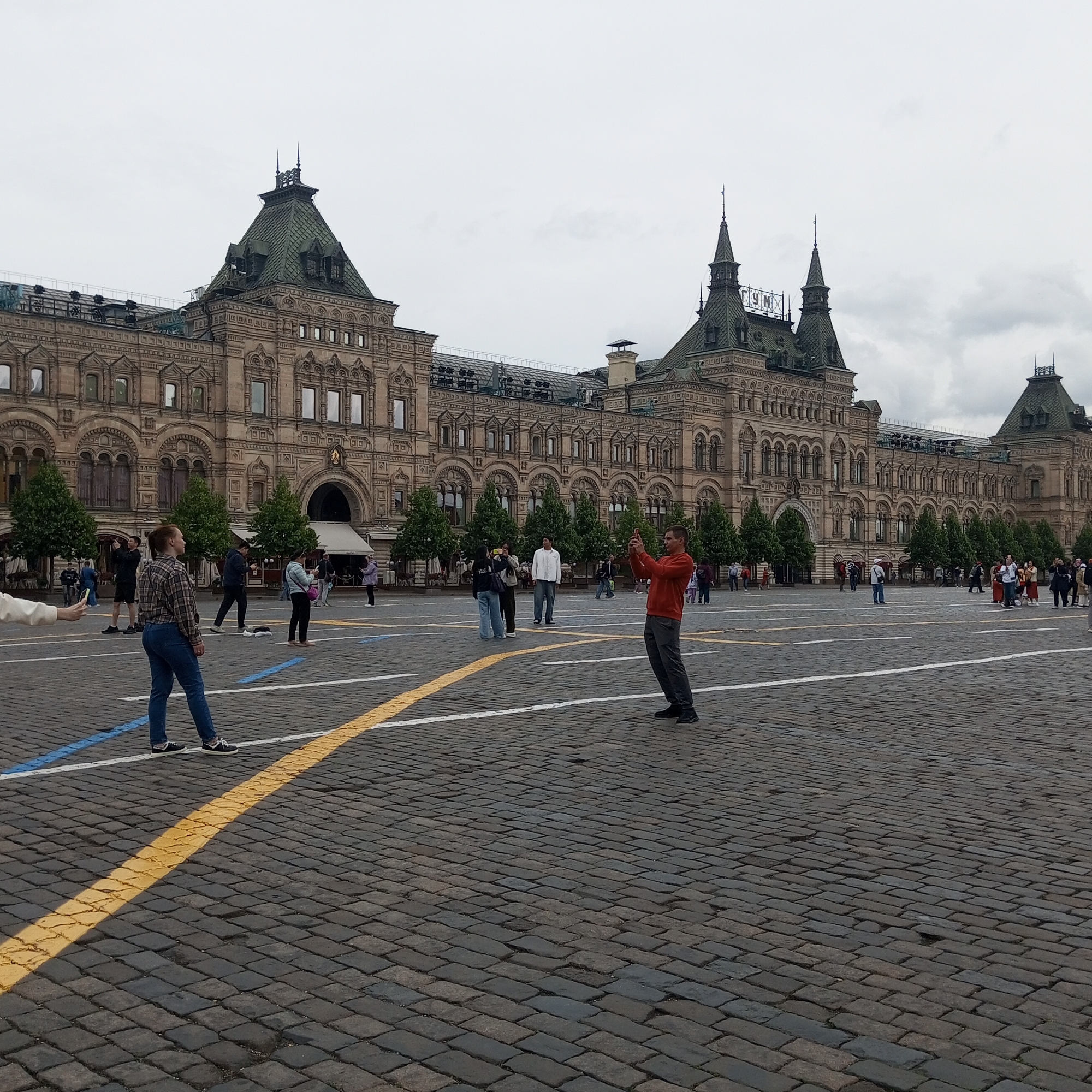 In the image, a man in an orange shirt is standing on a cobblestone plaza, holding a smartphone up to take a photo. He is facing a large, ornate building with a green, mansard roof and numerous windows. Behind him, a woman in jeans and a plaid shirt is also looking away from the camera. Other people are visible in the background, some taking photos, and others walking or standing. The sky is overcast. The scene appears to be a public space, likely a tourist attraction given the presence of people taking pictures of the building.