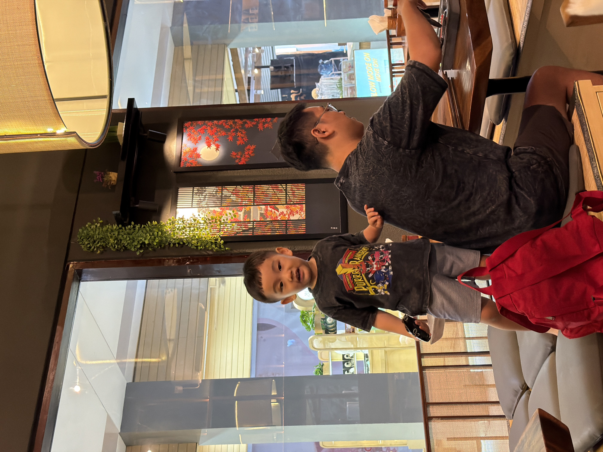 A vertical, eye-level shot captures two people in a dimly lit, indoor setting, possibly a restaurant or cafe. On the left, a young boy, appearing to be around four years old, grins at the camera. He wears a dark gray t-shirt featuring a graphic of the Power Rangers, along with light gray shorts. His left hand is positioned near his hip, while his right hand holds a dark object. To the right of the boy, an adult male is partially visible from the waist up. He is wearing glasses and a dark gray, textured t-shirt and is looking towards his right. His right arm is raised, holding a white object, possibly a napkin. Behind them, a bright red backpack is placed on a light-colored sofa.
The background reveals a large window or glass partition that reflects the interior and exterior. Through the glass, a Japanese-style painting with red maple leaves and a full moon can be seen on the wall. Further back, there are glimpses of other patrons and the restaurant's decor, including tables and seating. The lighting suggests it might be evening or that the interior is designed for a subdued ambiance. The overall mood is casual and relaxed. The location context provided is Kuta, Indonesia.