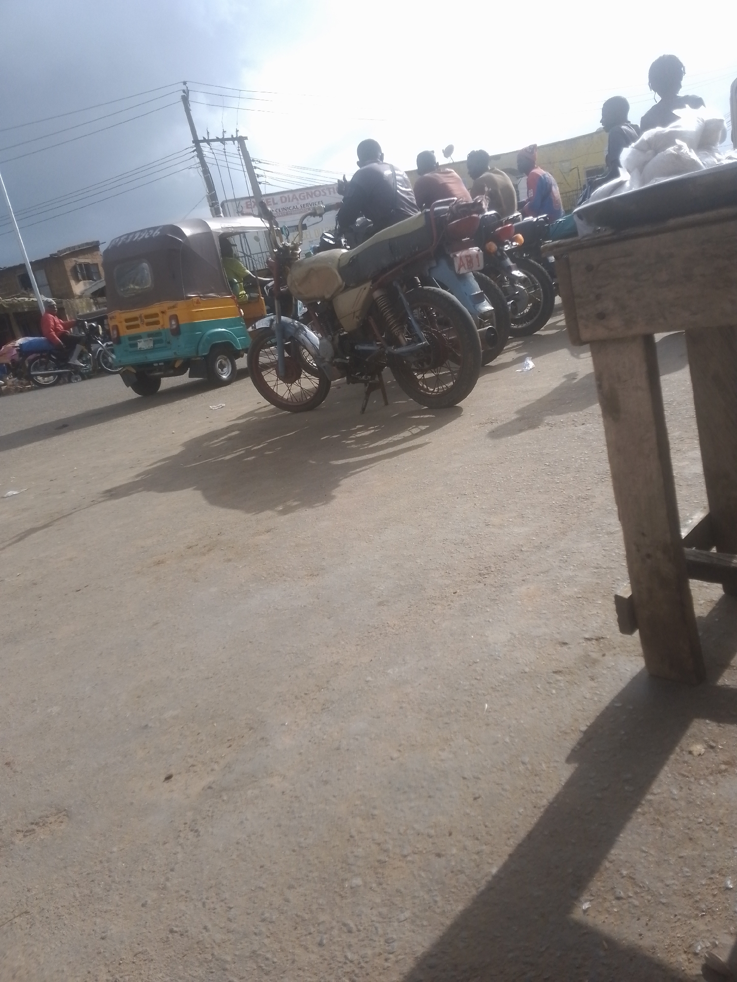 The image depicts an outdoor scene, likely a street or market, with several motorcycles and a tricycle. The sky is overcast. In the foreground, a wooden table or stand is partially visible on the right, casting a long shadow across the dusty ground. Several motorcycles are parked in a row behind the table. One motorcycle in the center, with a light-colored seat and frame, is more prominently featured. The tricycle, yellow and teal in color, is visible on the left side of the image, with a driver in a green shirt inside. Buildings with signage, including "EXCEL DIAGNOSTIC", are visible in the background, suggesting an urban or semi-urban setting. The overall impression is of a bustling area with transportation and commerce.