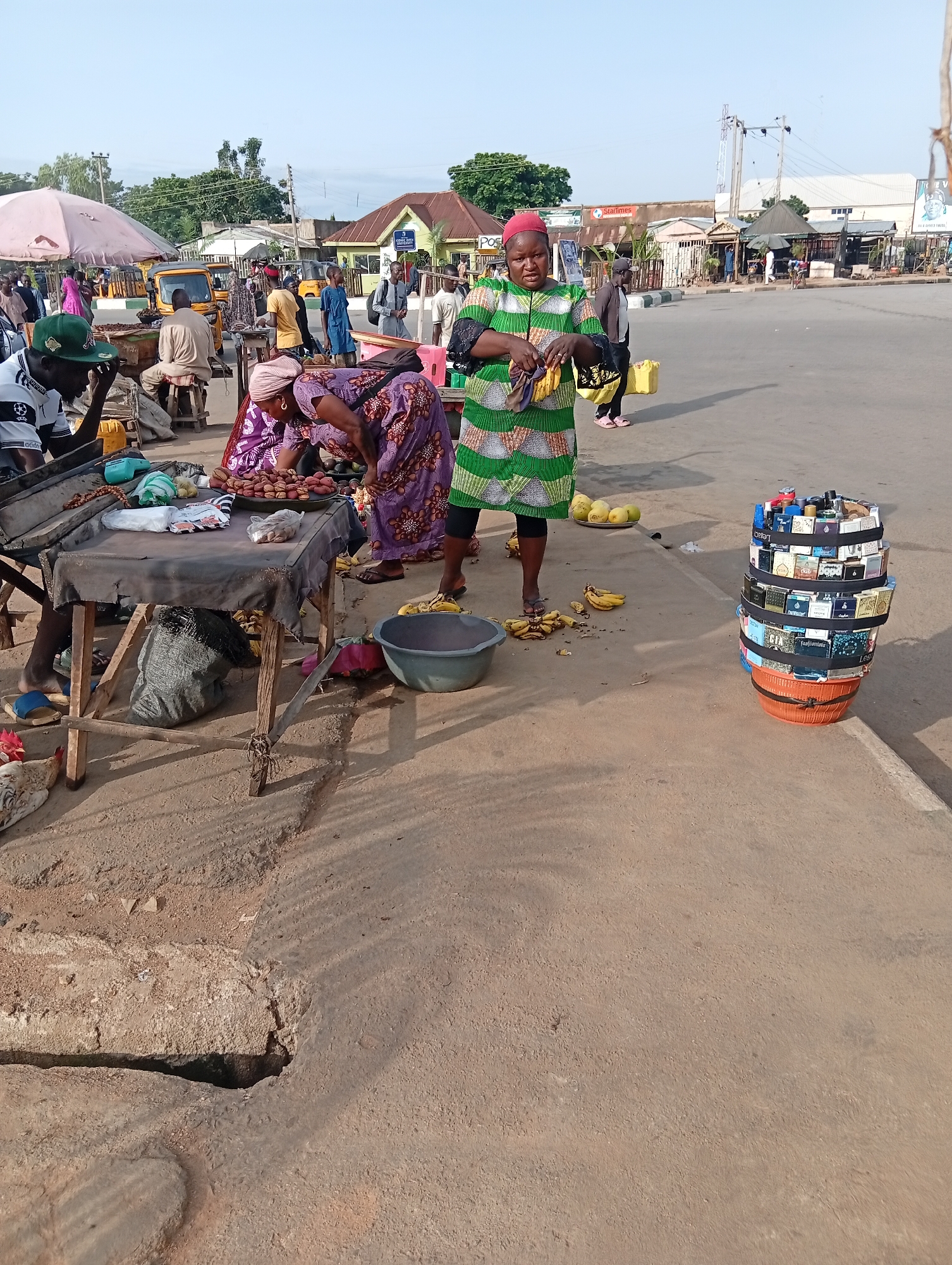 In Jalingo, Nigeria, an outdoor market scene unfolds under a bright sky. A woman in a green and white striped dress stands on the right, holding bananas. To her left, another woman in a purple patterned dress bends over a market stall laden with fruits and other goods. A man wearing a green cap and white shirt is seated at the stall, appearing to be a vendor. Behind them, various other market stalls and individuals are visible, along with a yellow three-wheeled vehicle. The ground is rough, dusty earth with a large pothole in the foreground. On the right, a tall, cylindrical display of perfume bottles is stacked in a basket. The background shows a street with more buildings and market structures.