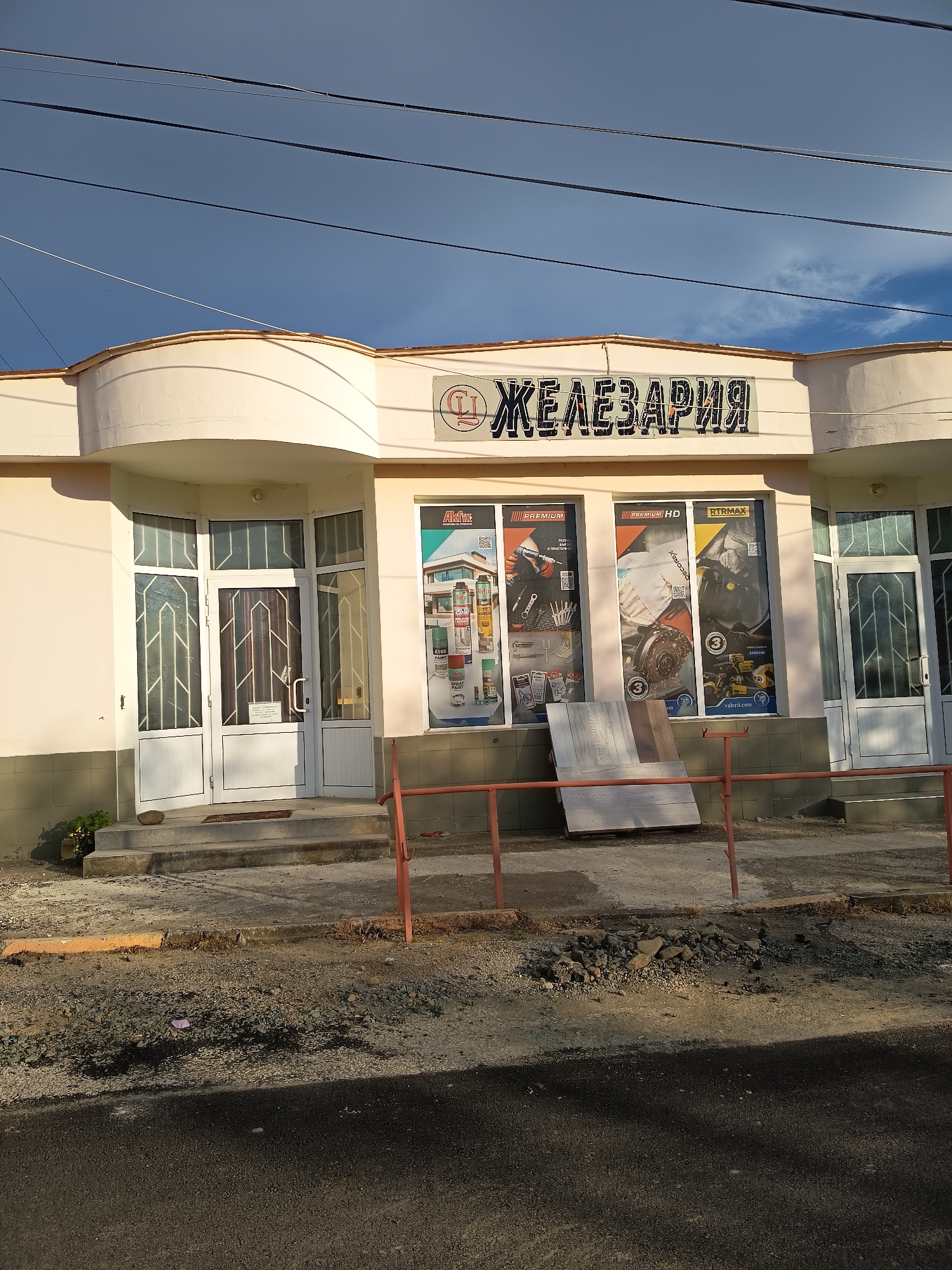 The image displays the exterior of a one-story building, likely a store, under a clear blue sky with some wispy clouds and utility wires crisscrossing overhead. The building has a light-colored, possibly beige or off-white, facade featuring curved architectural elements above its entrances.
The most prominent feature is the large sign on the building's front, which reads "ЖЕЛЕЗАРИЯ" in Cyrillic, translating to "Hardware Store" or "Ironware Store." Below the main sign, two large display windows flank the right entrance. These windows showcase advertisements for various products, including tools, automotive parts (like clutches and engine components), and spray paints. Visible brand names and text include "Auto," "Akfix," "PREMIUM HD," "RTRMAX," "SAMSUNG," and "valex.com." A small red circular logo, possibly containing the letters "СЦ" or "CU," is visible on the facade above the right entrance and on the building's left curve.
The building has two main entrances. The left entrance features a double door with decorative glass panels and a single step. The right entrance has a similar single door.
In the foreground, there is an asphalt road, leading up to a rough area of dirt, gravel, and small rocks. A low, reddish-brown metal railing separates this unpaved area from the storefront. Leaning against the railing near the right window are several flat, possibly wooden, boards. A small pile of rocks and some scattered litter, including a pink piece of plastic, are visible on the ground. A small green plant grows near the base of the left entrance.
The lighting suggests it is either morning or late afternoon, with long shadows cast by the utility wires onto the building's facade. No people are visible in the scene, and the overall impression is one of a quiet street.