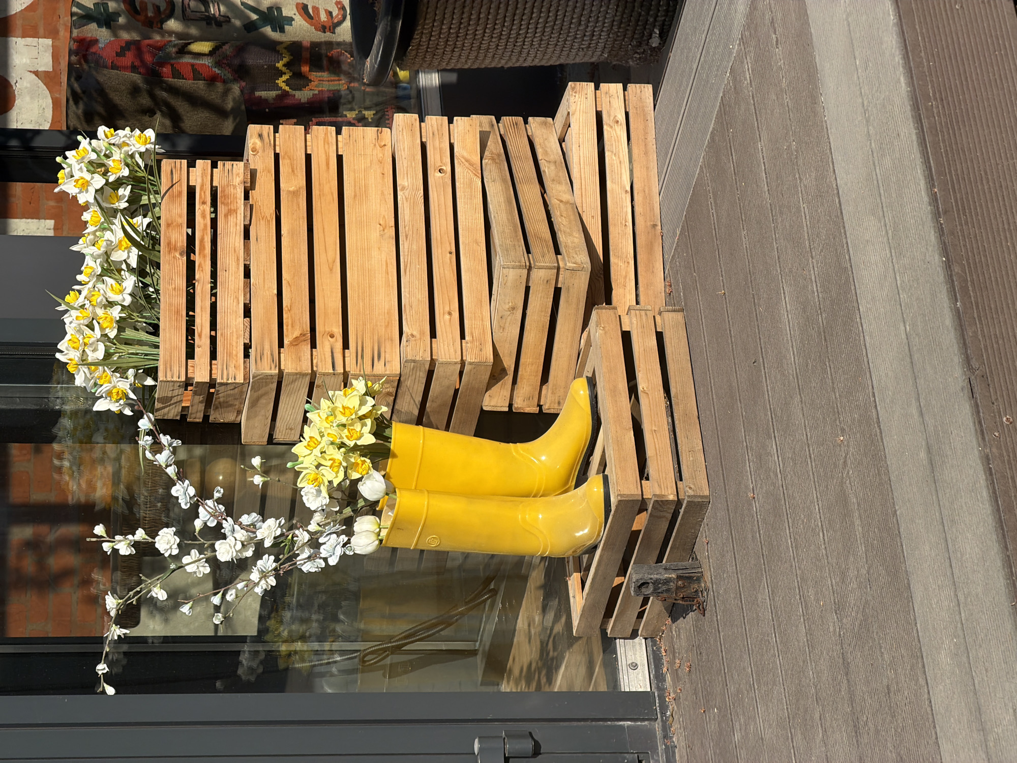 A street scene depicts a storefront display in Moscow, Russia. A wooden crate-style bench holds a pair of bright yellow rubber boots. The boots are filled with a bouquet of white and yellow flowers. The display is set against the storefront's window, which reflects the surrounding brick building and a patterned rug. The scene is outdoors, with a wooden deck visible on the right.