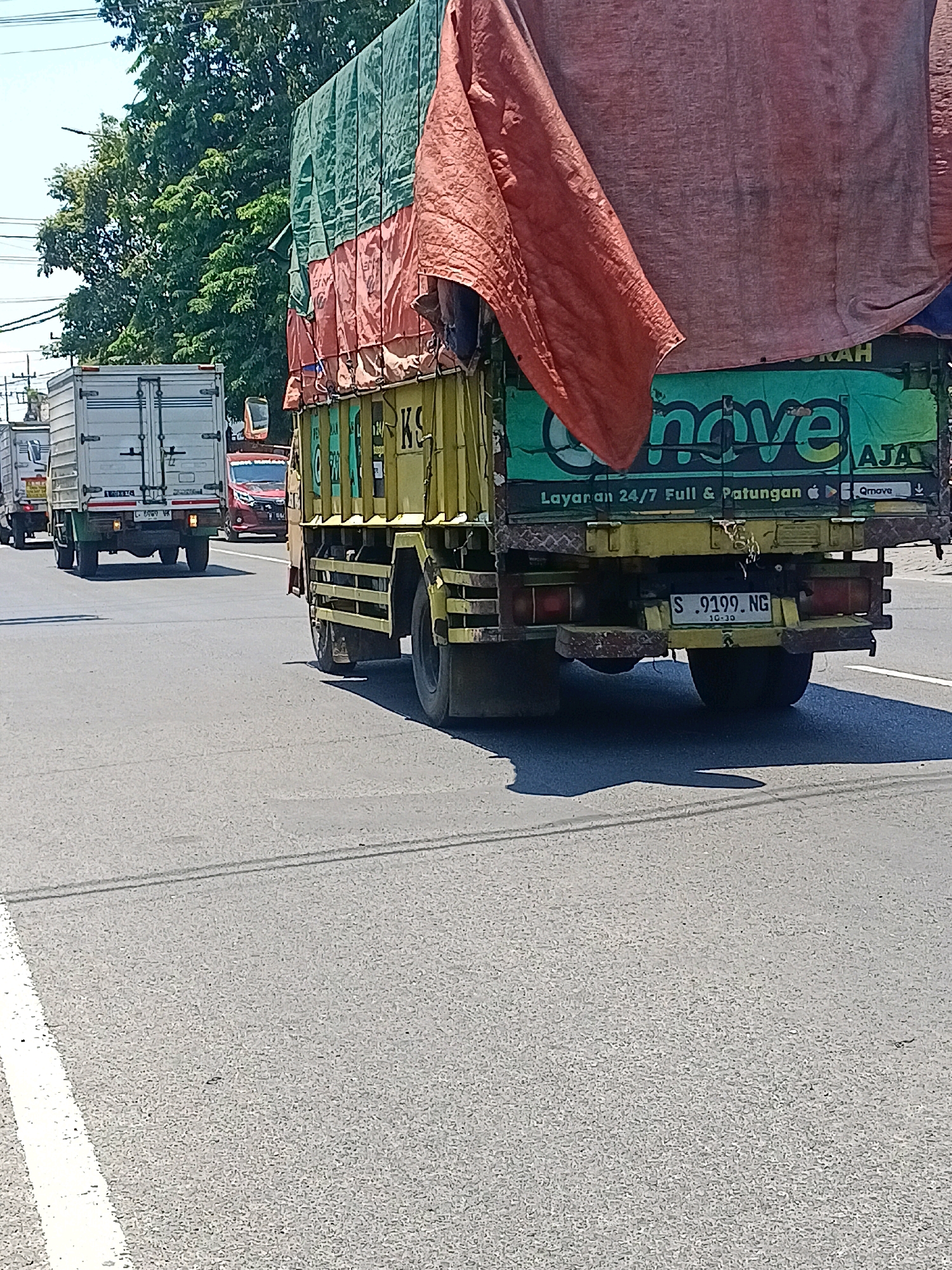 In Kalijaten, Indonesia, on a bright, sunny day, a line of trucks drives down a paved road. The foreground is dominated by the rear of a yellow and green truck, its cargo covered by a red and green tarp. The truck's license plate is visible, reading "S. 9199. NG". To the left, other trucks are visible in the middle ground and background, including a white truck with a silver cargo box. The road has white lane markings. Trees line the side of the road in the background, and a clear blue sky is overhead.