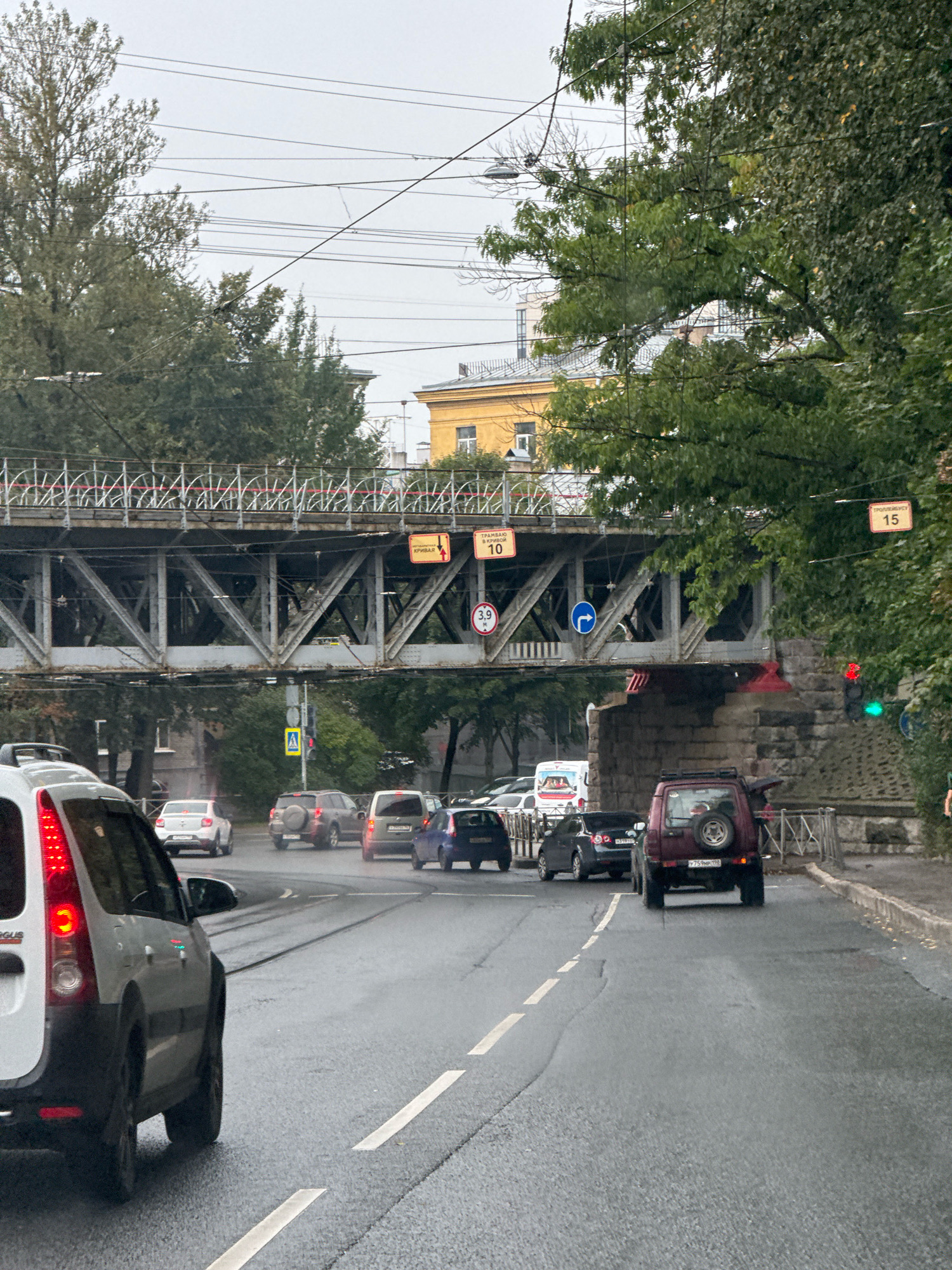 The image depicts a street scene in Saint Petersburg, Russia, under an overcast sky. In the foreground, a white vehicle is visible from the rear quarter, facing away from the camera and towards a traffic light. The road is wet, suggesting recent rain. Several other vehicles are driving or stopped on the road, including a dark red SUV in the middle lane.
Above the road, a metal pedestrian bridge crosses from left to right, supported by a lattice structure. Yellow road signs are attached to the bridge and nearby poles. One sign reads "ТРАМВАЙНАЯ ОГРАНИЧЕНИЯ 10" (Tramway Restriction 10), another displays "3,9" with a circle around it, and a third sign indicates "15". A blue directional arrow sign is also visible.
To the right of the road, a stone embankment is visible, and further back, trees with green foliage line the street. Buildings with yellow and lighter facades are visible behind the trees. Power lines crisscross the sky. A traffic light shows a red light on top and a green light below. The overall impression is of an urban environment during daytime, likely in the autumn season due to the leaves on the trees.