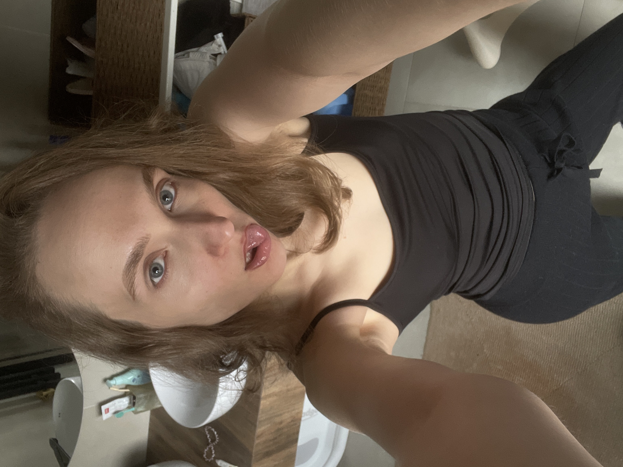 A young woman is captured in a high-angle selfie, rotated 90 degrees counter-clockwise. She has light skin, medium-length brown hair, and striking blue eyes. Her gaze is directed towards the camera with a wide-eyed expression, and her lips are slightly parted, revealing her tongue in a playful manner. She is wearing a simple black tank top and black ribbed pants.
The setting appears to be an indoor, private space, likely a bathroom or dressing area in Podshivalovo, Russia. In the lower left portion of the rotated image, a white sink and a portion of a vanity counter are visible. On the counter, there's a small white bowl or dish, and various small objects, including one with illegible red text. The background features a wooden shelf unit and a light-colored, possibly white, decorative object. The floor, visible in parts, appears to be a light-colored tile or carpet. The lighting is ambient indoor light.