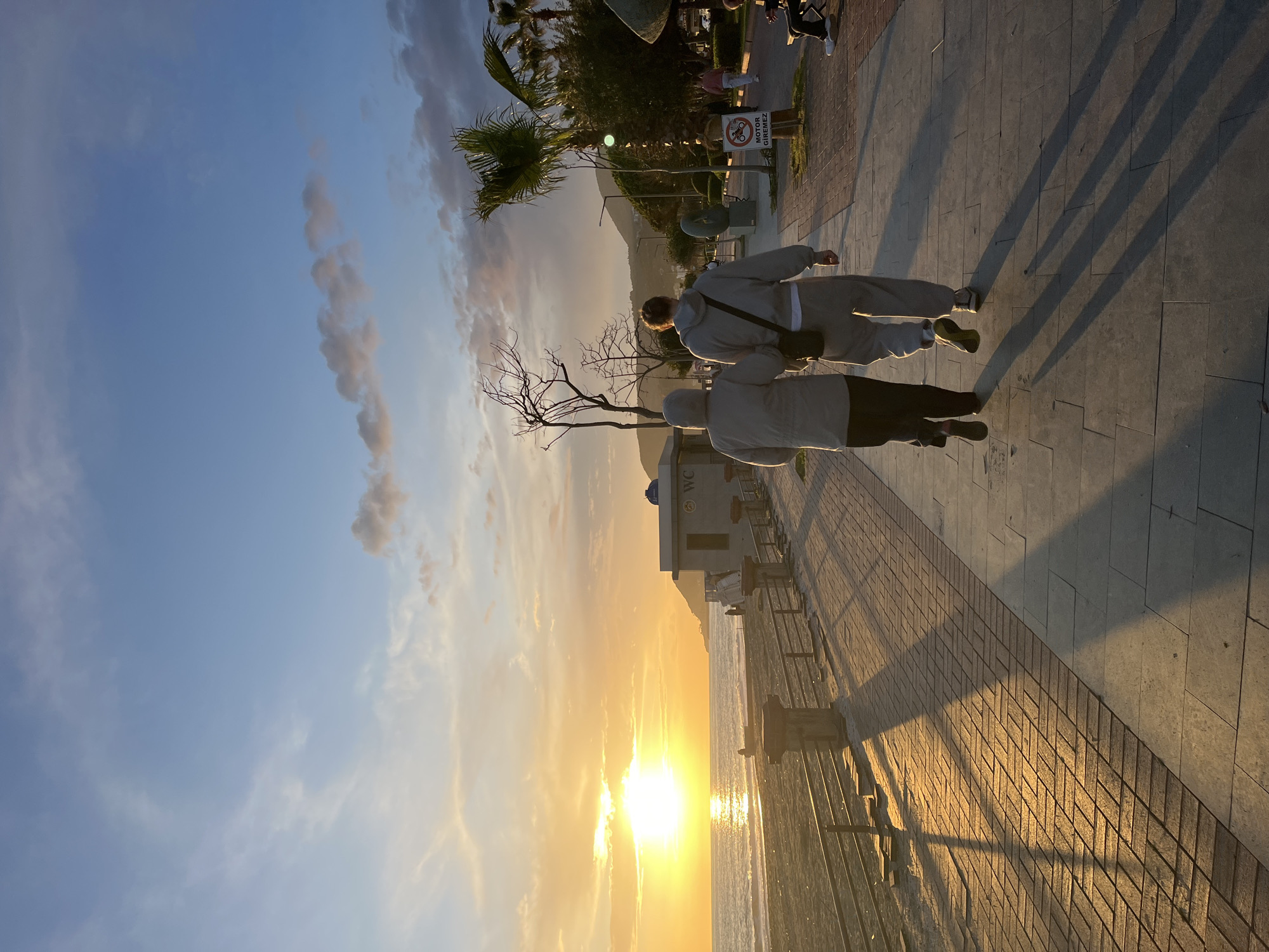 The image shows a seaside scene at sunset in Alanya, Turkey. The sun is setting over the water, casting a golden light across the scene. Two individuals are walking along a paved pathway parallel to the beach. The sky transitions from a light blue to a vibrant orange near the horizon. Palm trees and other greenery are visible along the walkway, as well as a sign prohibiting motorcycles. There are building structures and bare trees along the path. The pathway is made of rectangular pavers, and railings separate it from the sandy beach and sea. Shadows are long and pronounced due to the low angle of the sun.