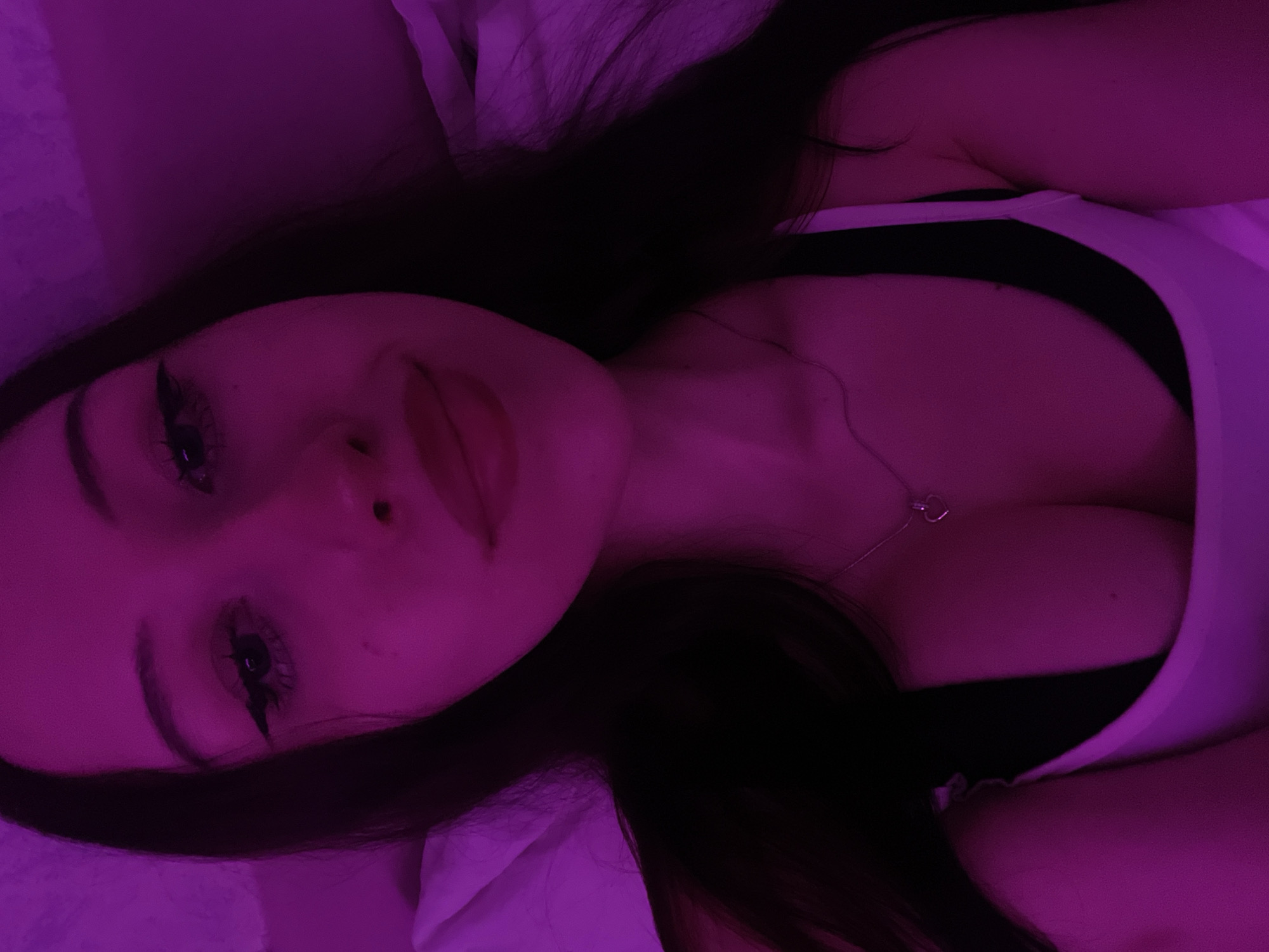 The image is a close-up, selfie-style photograph of a young woman with long, dark hair, illuminated by purple lighting. She is looking directly at the camera, with heavy eyeliner and lipstick. She is wearing a white tank top over a black bra, and a delicate necklace with a heart-shaped pendant. The background is softly blurred, suggesting an indoor setting, possibly a bedroom. No specific location in Votkinsk, Russia, can be determined from the image.