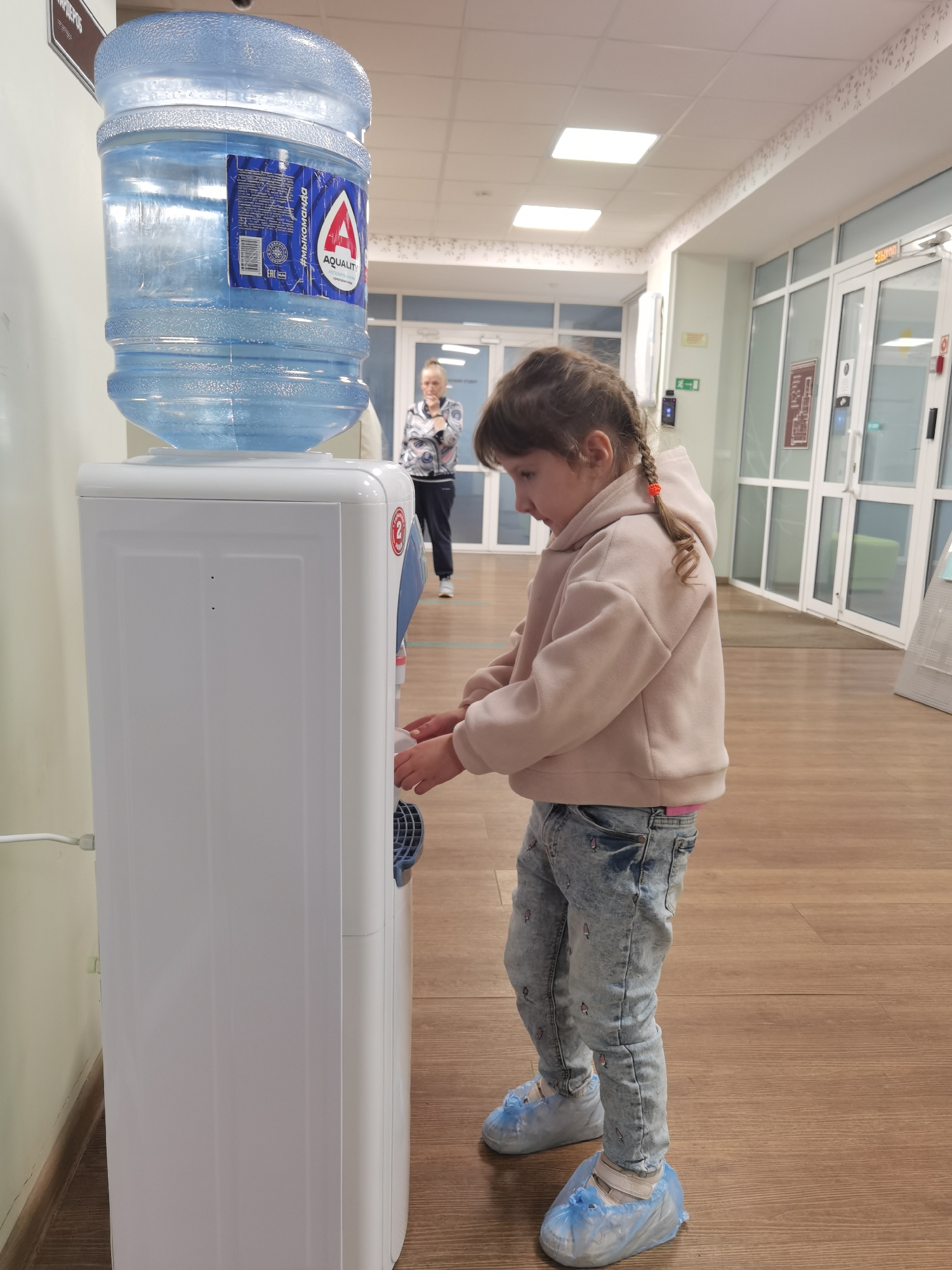 An indoor scene in a modern corridor of a medical facility, identified as Doctor Karyerov's Clinic (Клиника доктора Карьерова) in Izhevsk, Russia. A young girl, positioned on the right, with light brown hair styled in two small braids, is wearing a light pink hooded fleece sweatshirt, acid-wash jeans with subtle patterns, and blue disposable shoe covers over white sneakers. She is actively filling a small white cup with water from a white, floor-standing water cooler on the left. The cooler features a large, inverted 19-liter bottle of "AQUALIT" brand water, which has a blue label displaying a large "A" logo and the Cyrillic text "#мыкоманда". A red circular emblem with the number '2' is visible on the cooler's right side. In the background, further down the corridor, an adult woman with light hair tied back, wearing a black and white patterned top and dark pants, stands observing. The corridor has light green walls with a wooden baseboard and light brown wooden flooring. The white ceiling is fitted with square LED panel lights. Along the right side, multiple floor-to-ceiling glass doors with white frames lead to other rooms. A red "ВЫХОД" (EXIT) sign is affixed above one of these doors, and a green emergency exit sign (running man icon) is visible on the left wall. A brown sign with the partial Cyrillic text "КАРЬЕРОВ" hangs high on the left wall.