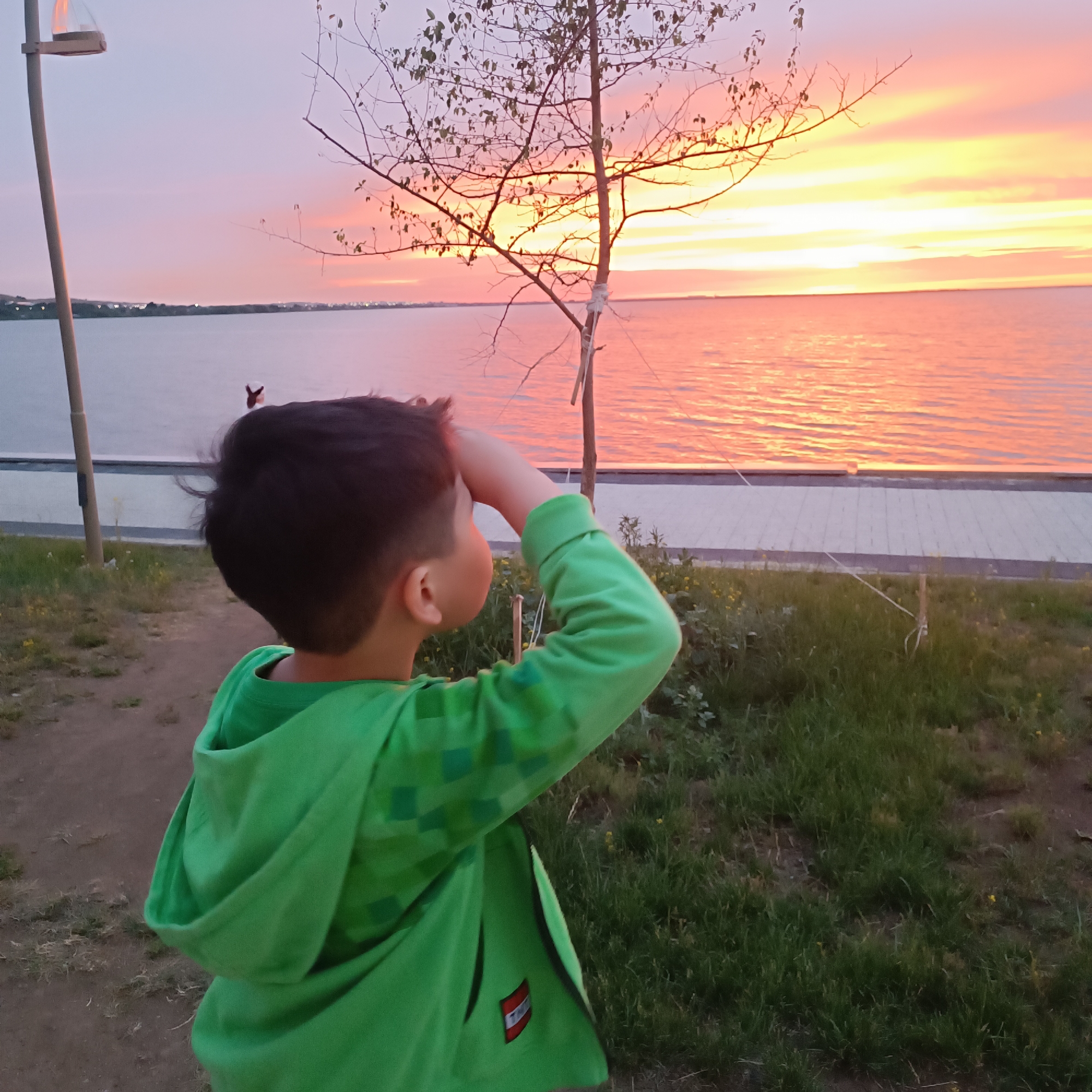 A young boy, seen from behind and in profile, stands on a grassy area looking out at a sunset over a body of water. He is wearing a bright green hooded sweatshirt with a pixelated pattern on the sleeves and a red and black logo on the front. The boy has his right hand raised to shield his eyes as he gazes towards the horizon. The sky is a vibrant display of pink, orange, and yellow hues as the sun sets. The water reflects the colors of the sky, creating a warm and inviting scene. In the background, a thin tree with sparse leaves stands to the right of the boy. A paved walkway runs along the edge of the water, and beyond that, the distant shoreline is visible with faint lights suggesting a town or settlement. A tall pole with a torch-like fixture is visible on the left side of the image. The scene is set in the evening, with soft lighting characteristic of twilight. The overall mood is one of peaceful observation and wonder. The location is Kokshetau, Kazakhstan.