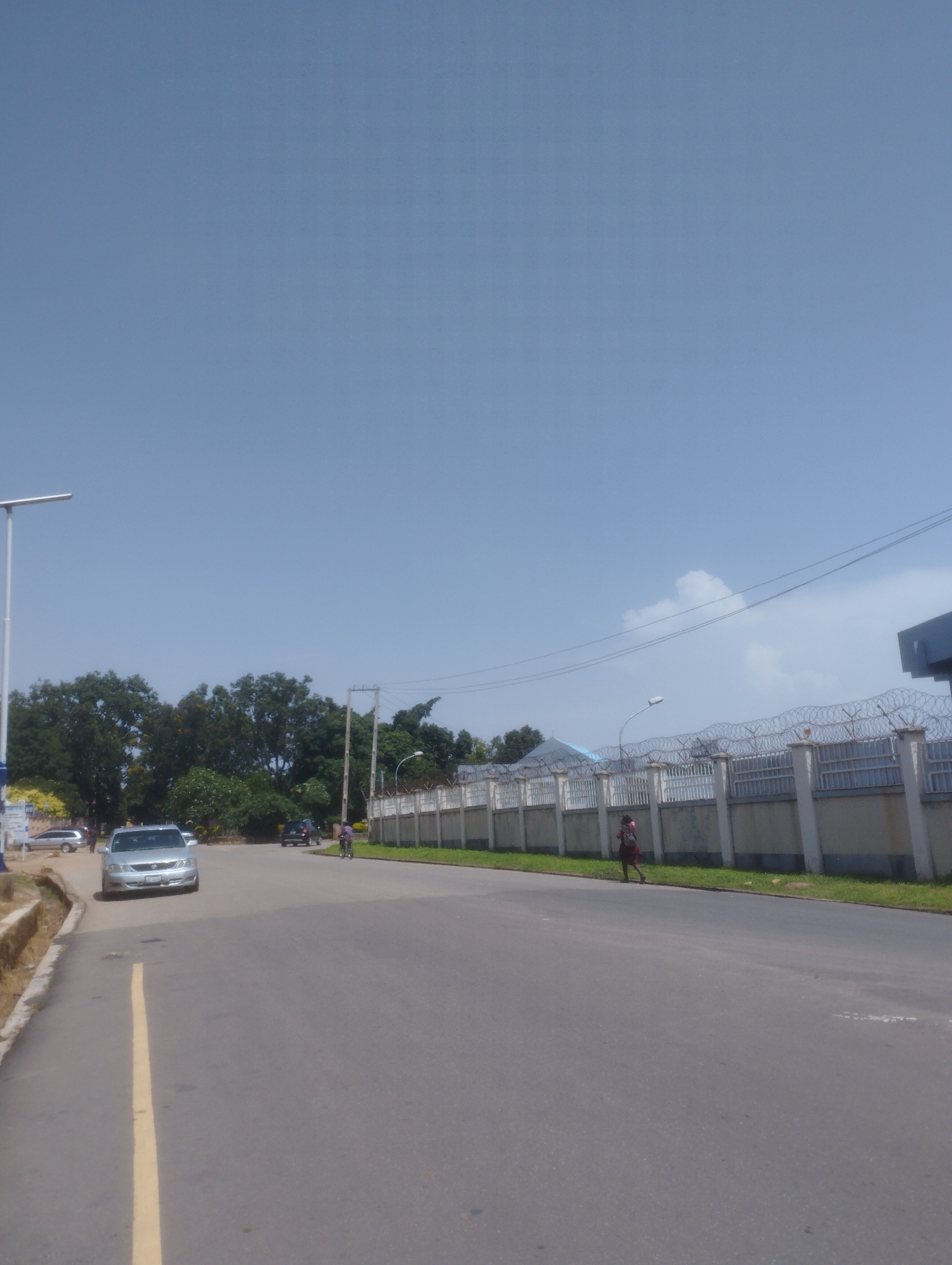 The image depicts a street scene in Jos, Nigeria, under a clear blue sky. A paved road runs diagonally from the lower left to the upper right of the frame. On the left side of the road, a raised concrete embankment lined with a yellow stripe is visible. Further left, a silver sedan is parked. A modern, white street lamp stands on the far left.
The right side of the road features a long, light-colored concrete wall topped with barbed wire, indicating a secure perimeter. Beyond the wall, a building with a dark blue roof is partially visible. A few trees are present in the background, with denser foliage on the left side of the scene.
Several vehicles are on the road: a silver sedan in the foreground on the left, and a dark-colored SUV further down the road on the right. A person on a bicycle is also visible on the right side of the road, moving away from the viewer. A person, possibly a woman with a backpack, is walking on the grassy area between the road and the wall on the right. Overhead, power lines are strung across the sky. The overall impression is a moderately busy urban street with some greenery and clear weather.