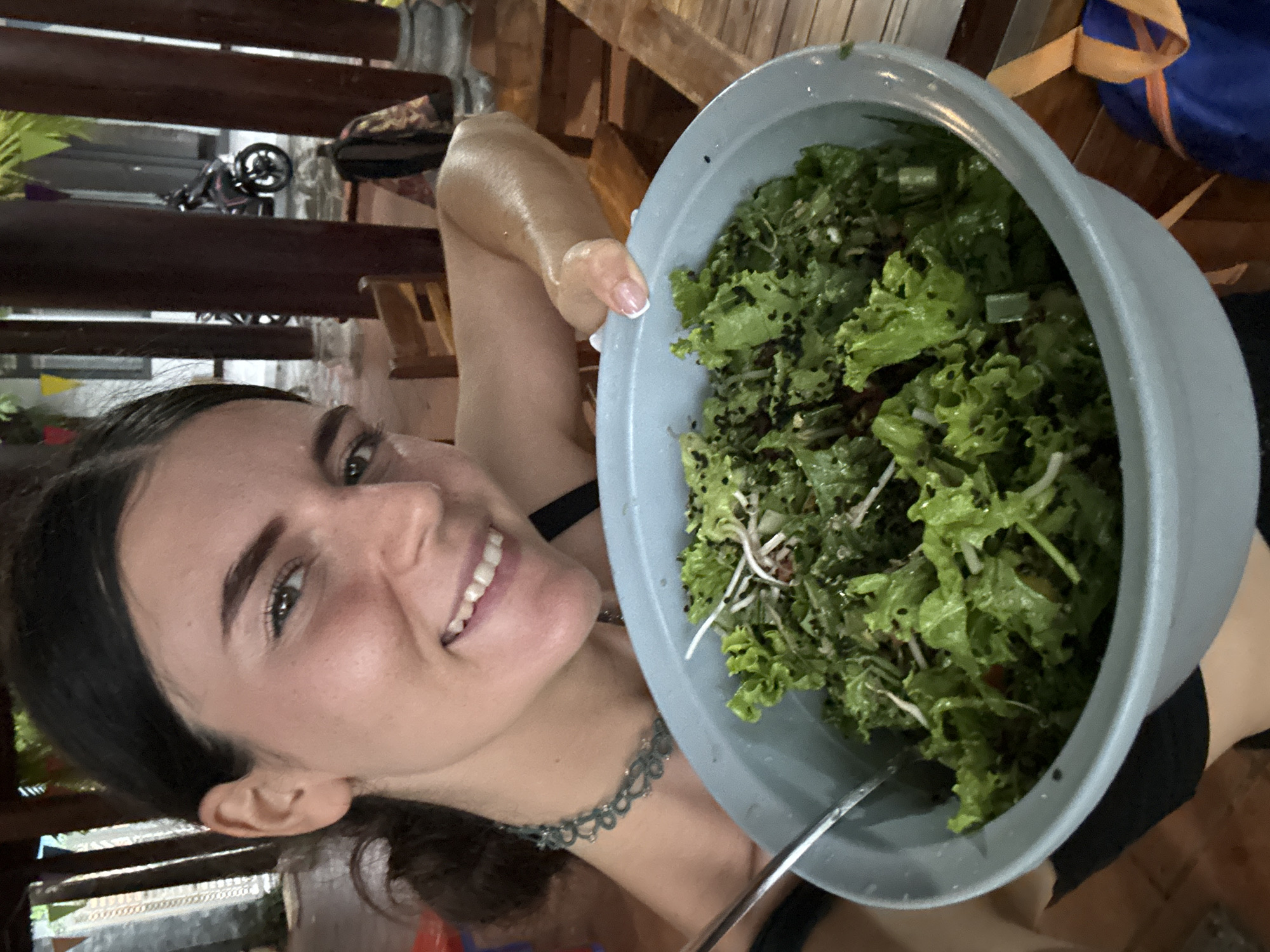 A woman is holding a large bowl of salad in front of her. The salad appears to contain green lettuce, sprouts, and other ingredients. She is smiling at the camera. She is wearing a dark top and a necklace. The background shows a wooden structure, possibly a porch or deck, and glimpses of other objects including a bicycle and luggage. The location is possibly in Mũi Né, Vietnam.