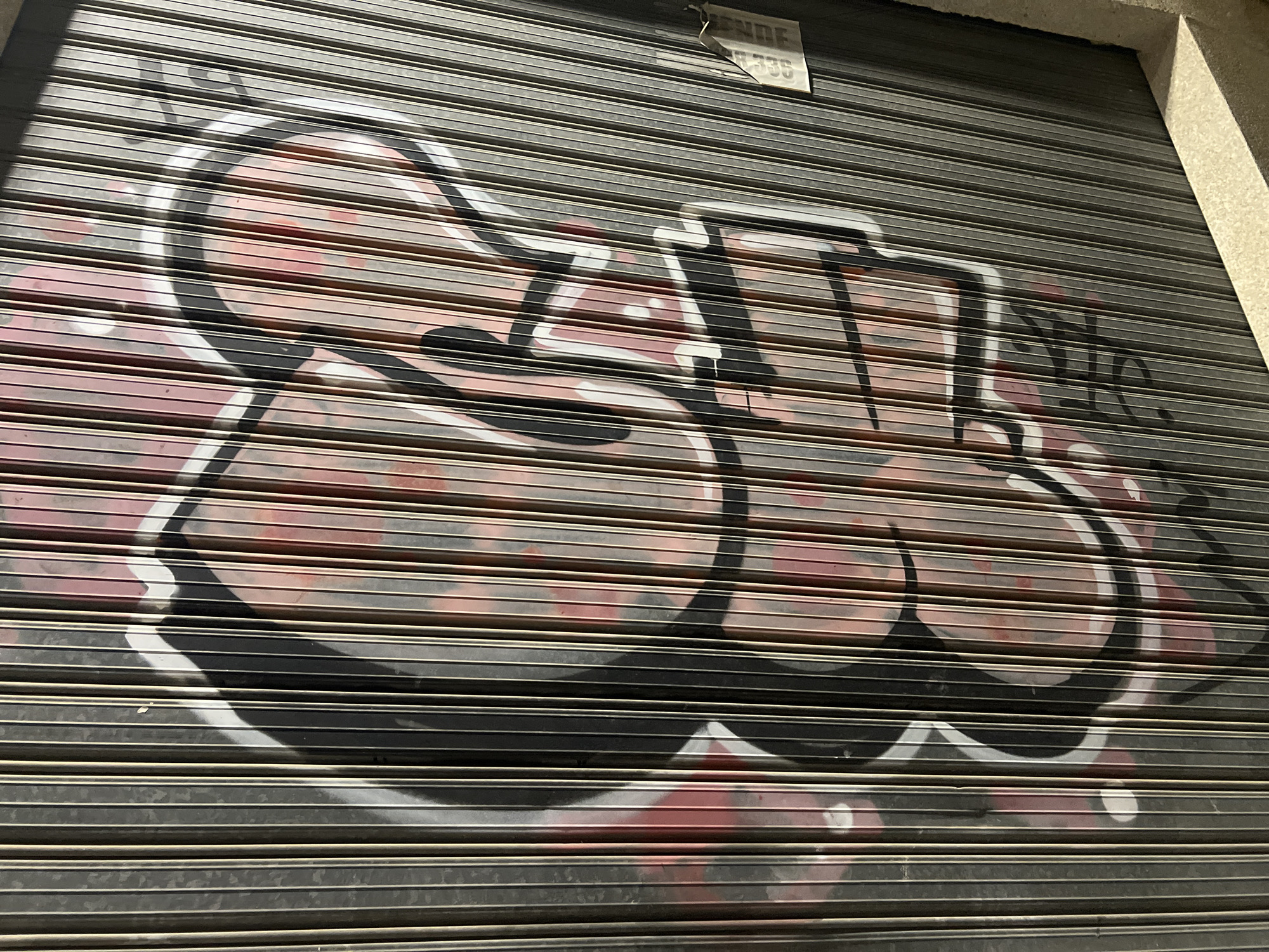 A corrugated metallic roller shutter, securing a building facade, is depicted in Picassent, Spain. The shutter is uniformly grey with horizontal interlocking slats. The dominant feature is a large, stylized graffiti tag rendered in bubble letters. This graffiti features thick black outlines, an interior fill gradient transitioning from deep red to lighter pinks, and white highlights providing a three-dimensional effect. The central portion of the graffiti clearly spells "SAKA" or "SANA," with a stylized 'S' followed by an 'A', a central letter resembling a 'K' or 'N', and a final 'A'. A small white paint drip is visible below the central letter. To the right of the main graffiti, a smaller, less distinct black tag is present. In the upper right quadrant of the shutter, a small, rectangular, light-colored sign is affixed, displaying partially legible dark text that includes the word "VENDE" (Spanish for "FOR SALE") and numerical digits, possibly "935" or "835". The scene is illuminated by ambient light, creating reflections on the metallic surface. No individuals are present in the image.