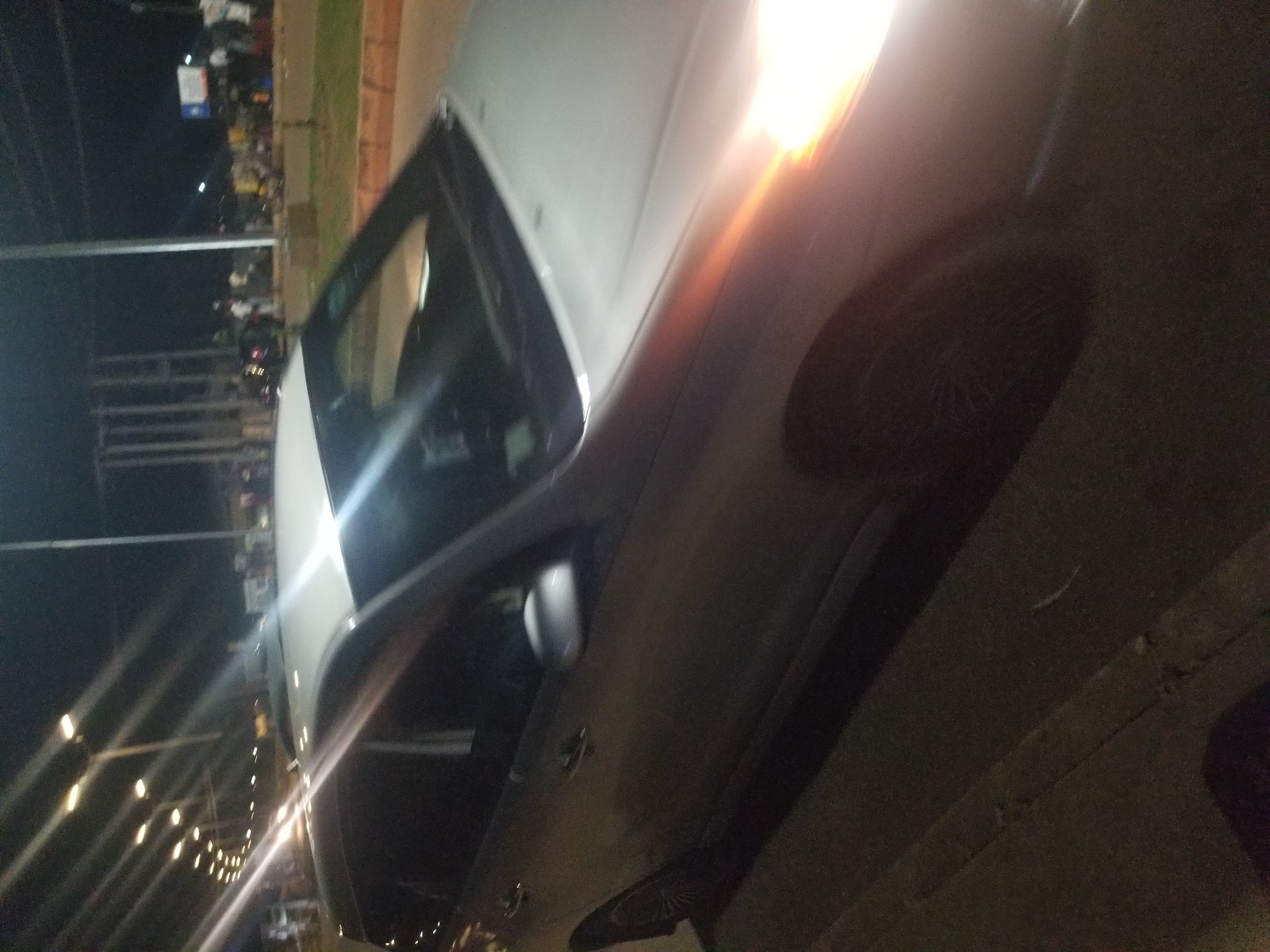 The image shows a silver station wagon parked on the side of a road at night. Streetlights illuminate the scene, casting long shadows. In the background, there is a group of people and vehicles, suggesting a busy area in Jos, Nigeria. The car's headlights are on, reflecting brightly on the vehicle and the road. The environment appears to be an urban setting with paved roads and sidewalks.