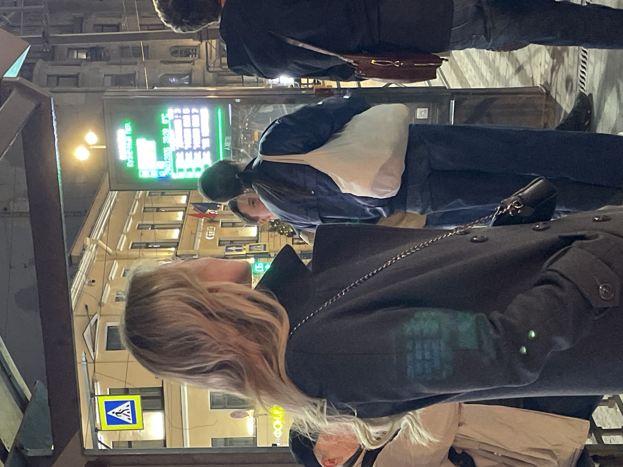 The scene shows a group of pedestrians on a street in Saint Petersburg, Russia, at night.
A blonde woman in a gray coat with a chain shoulder strap is in the foreground. Her back is to the camera. A woman with a black mask, light-colored hair, and a beige jacket is in front of her.
A woman with dark hair wearing a blue jacket and a white tote bag is in the middle ground. She is looking to the left. A man in a dark coat walks past.
In the background are buildings with lit windows. A green digital sign shows the weather. A pedestrian crossing sign is visible. A street lamp illuminates the scene.