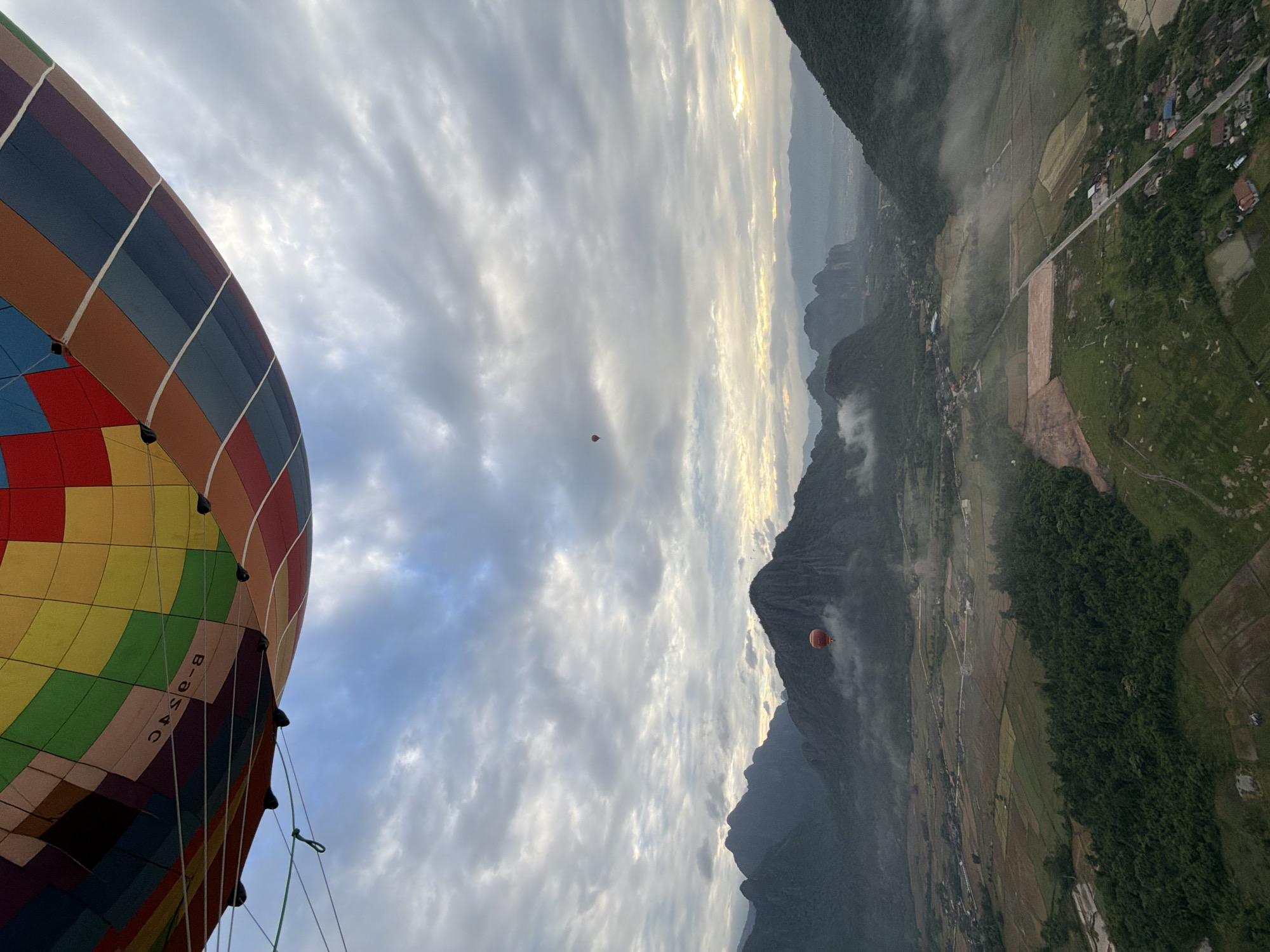 This media file captures a scenic view from a hot air balloon, likely during a sunrise or sunset given the soft, diffused light and the golden glow on the horizon. The scene is set in a mountainous region, possibly in Hong Ngern Village, Laos, as indicated by the presence of lush green hills, terraced fields, and a winding road.
The dominant subject in the foreground is a large, vibrantly colored hot air balloon, its patterned fabric a patchwork of reds, oranges, yellows, blues, and greens. The balloon is tethered and likely in the process of ascent or descent. In the distance, two smaller, red hot air balloons are visible, adding to the sense of wonder and adventure.
The landscape unfolds below, showcasing a rural setting with patches of cultivated land, verdant forests clinging to the slopes of the mountains, and a scattering of houses and buildings that suggest a small village. Wisps of mist or low-lying clouds drift across the valleys, adding a mystical quality to the panorama. The sky is filled with dramatic, textured clouds, some of which are illuminated by the sun's rays, creating a sense of depth and vastness.
The overall mood is one of peace and awe, with the expansive natural beauty and the gentle presence of hot air balloons evoking a feeling of tranquility and escape. There is no visible text other than some lettering on the large balloon, which appears to read "B-U-SAC". The time of day appears to be early morning or late evening due to the quality of light and the presence of clouds.