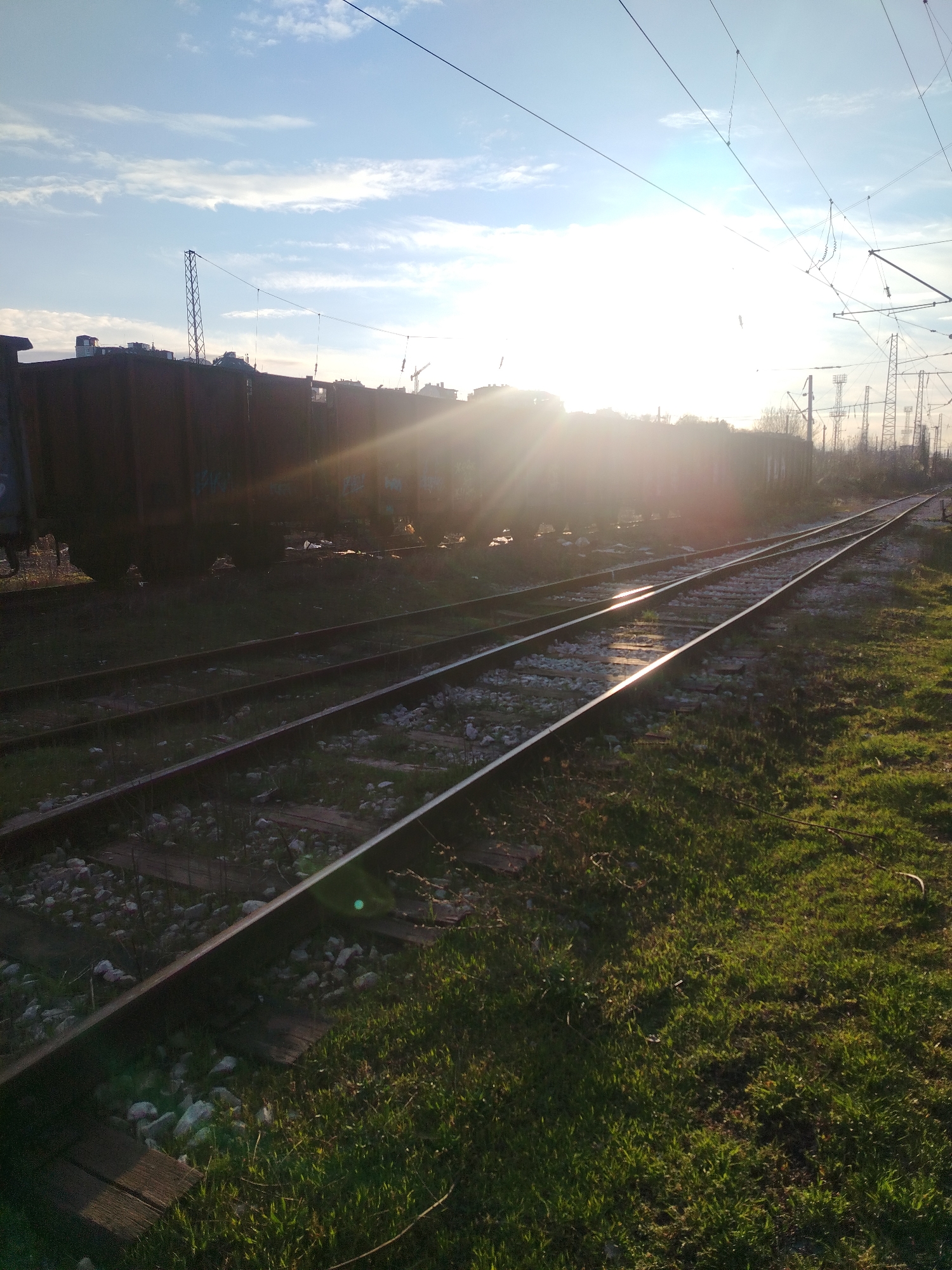 The image shows a section of railway tracks in Sofia, Bulgaria. The tracks run diagonally across the frame, with the right side of the image showing green grass along the tracks, and the left side having railroad cars parked on the tracks. The sun is directly in front of the train, almost obscuring the view. The sky is bright blue with a few scattered clouds. Overhead electrical lines and towers are visible in the background.