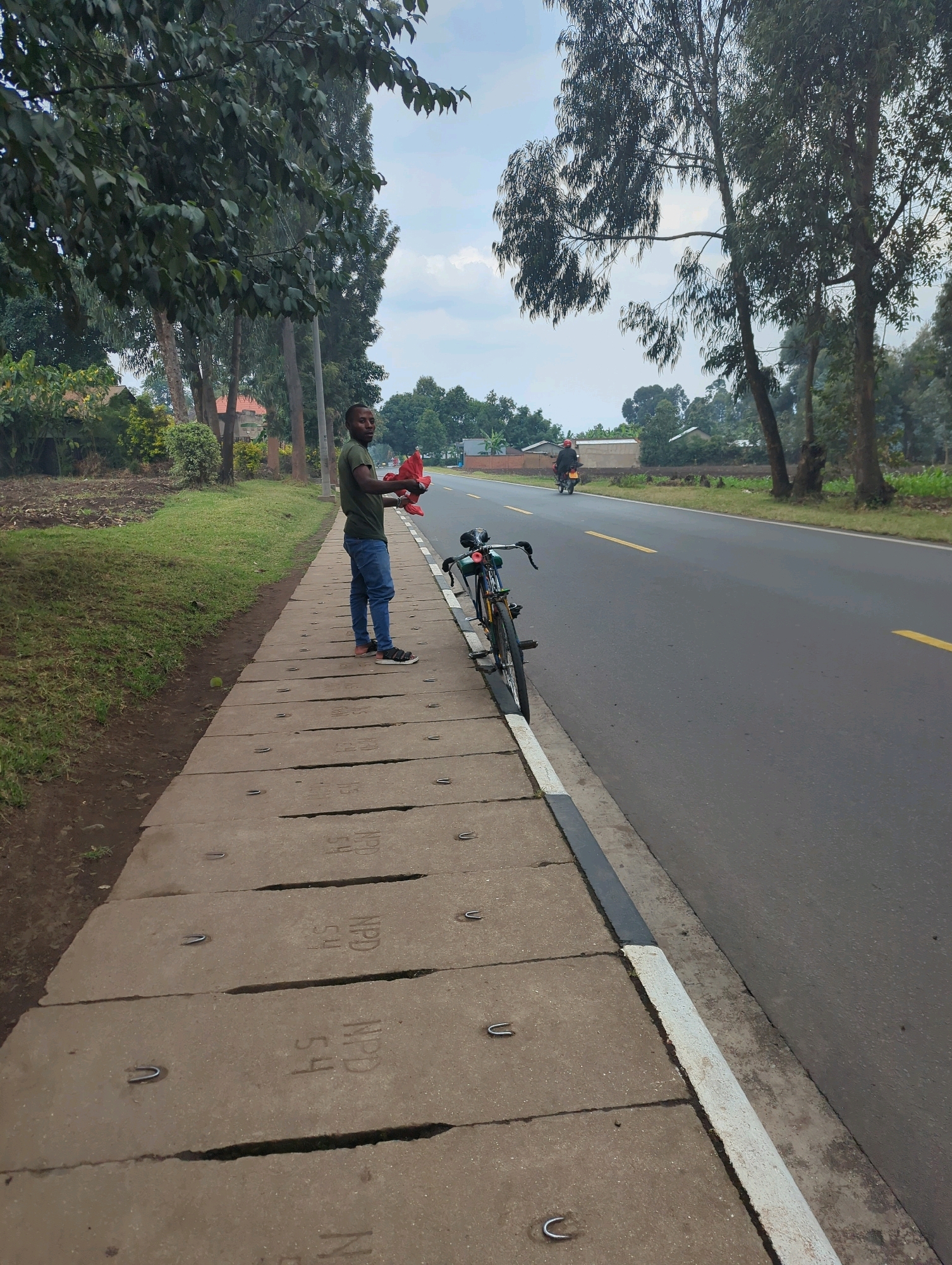 A man stands on a concrete sidewalk next to a paved road. He is wearing a green t-shirt and blue jeans, and is holding a red cloth in his left hand. A bicycle is parked next to him on the sidewalk. In the background, there are trees, houses, and a motorcycle driving on the road. The sky is overcast. The scene appears to be in a semi-rural or suburban area. The sidewalk has several pre-cast concrete slabs with metal loops. The man is looking towards the camera.